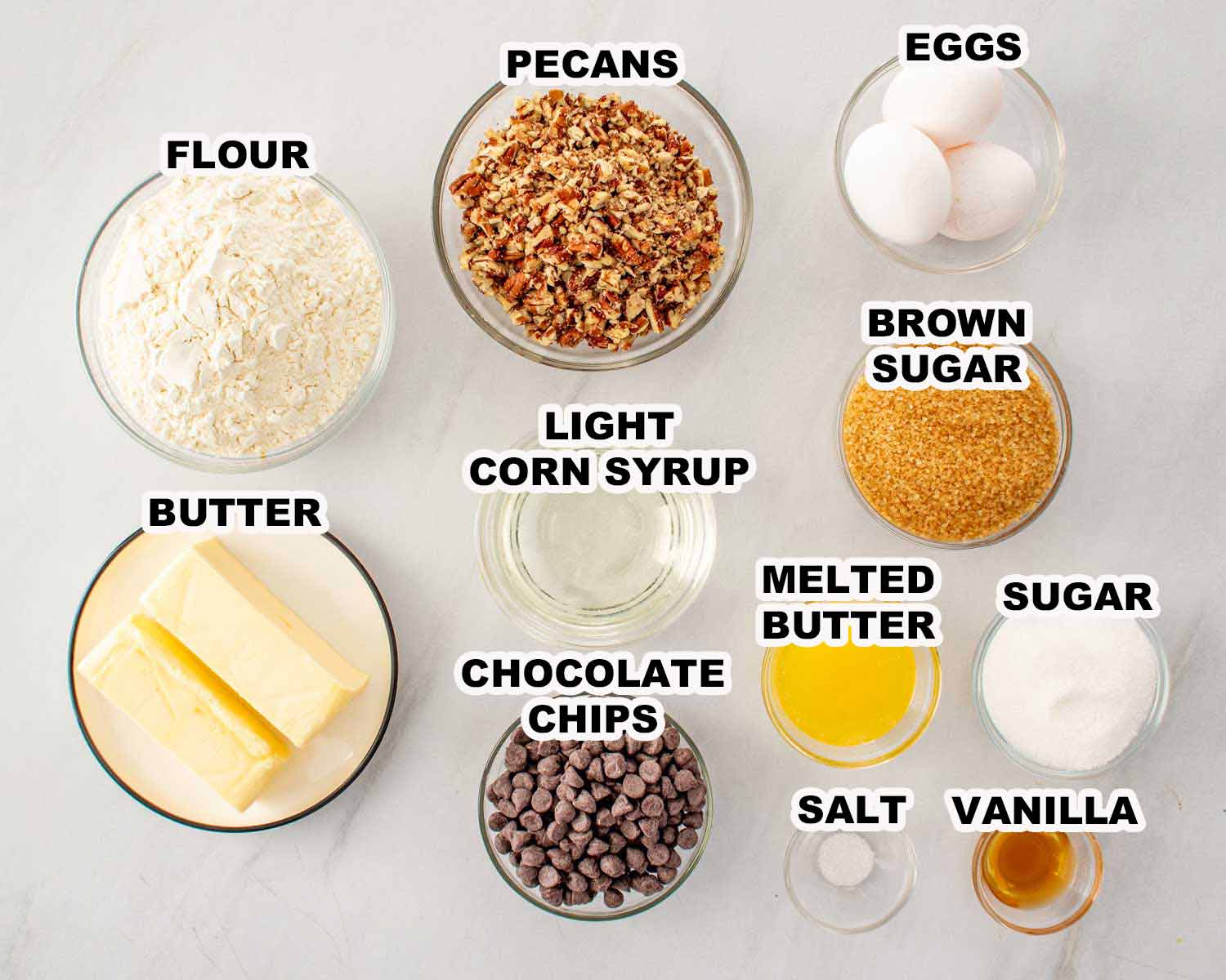 Flat lay of chocolate chip pecan pie bar ingredients including chopped pecans, eggs, flour, butter, light corn syrup, brown sugar, chocolate chips, melted butter, sugar, salt, and vanilla, each labeled on a white marble surface.
