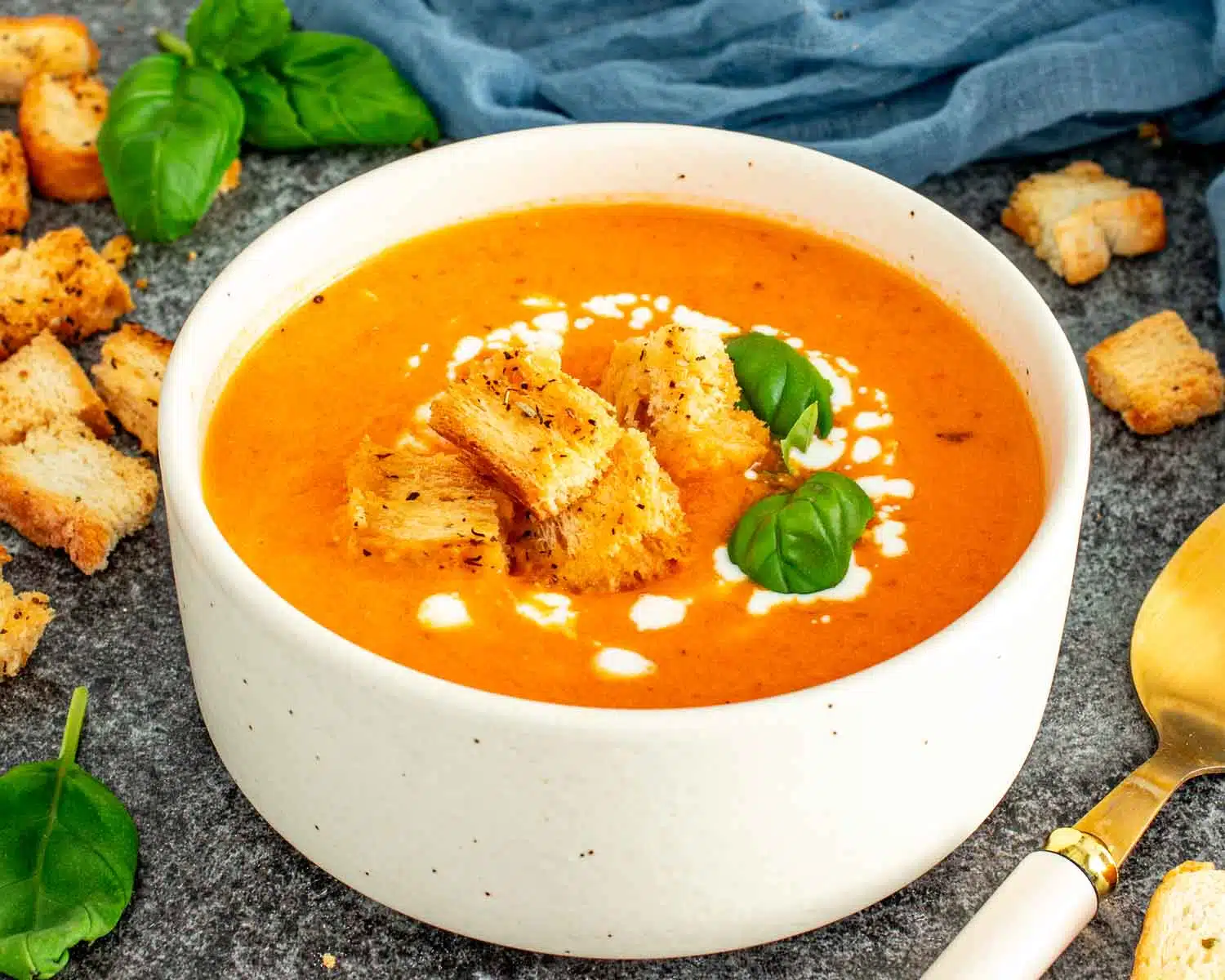 A white speckled bowl of creamy tomato soup topped with golden croutons, a cream drizzle, and fresh basil with a blue linen and gold spoon in the background.