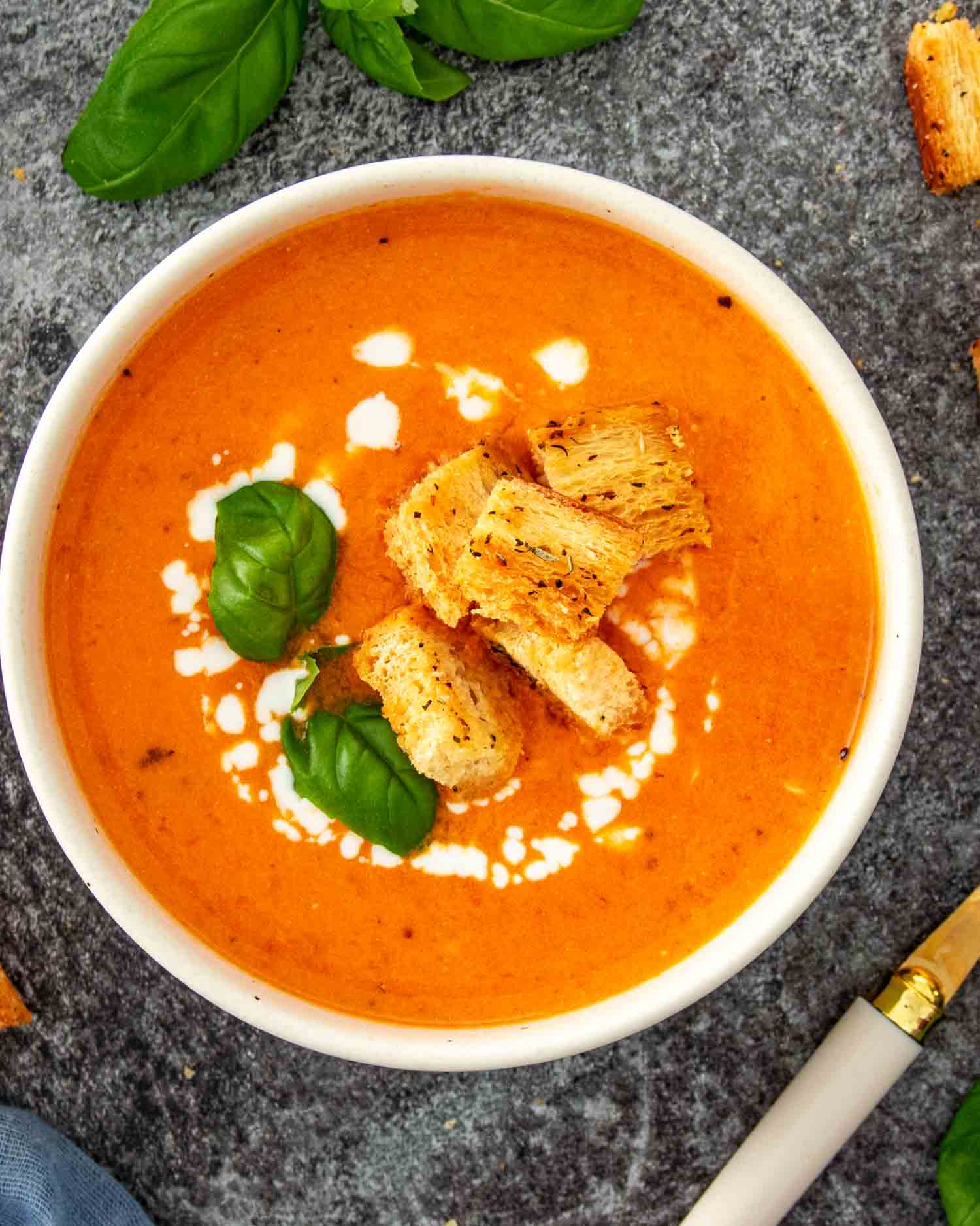 Overhead view of a white bowl of creamy tomato soup garnished with a cream swirl, golden croutons, and fresh basil leaves.