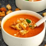 2 white speckled bowls of creamy tomato soup topped with golden croutons and fresh basil with a gold and white spoon resting in the front bowl surrounded by scattered croutons.