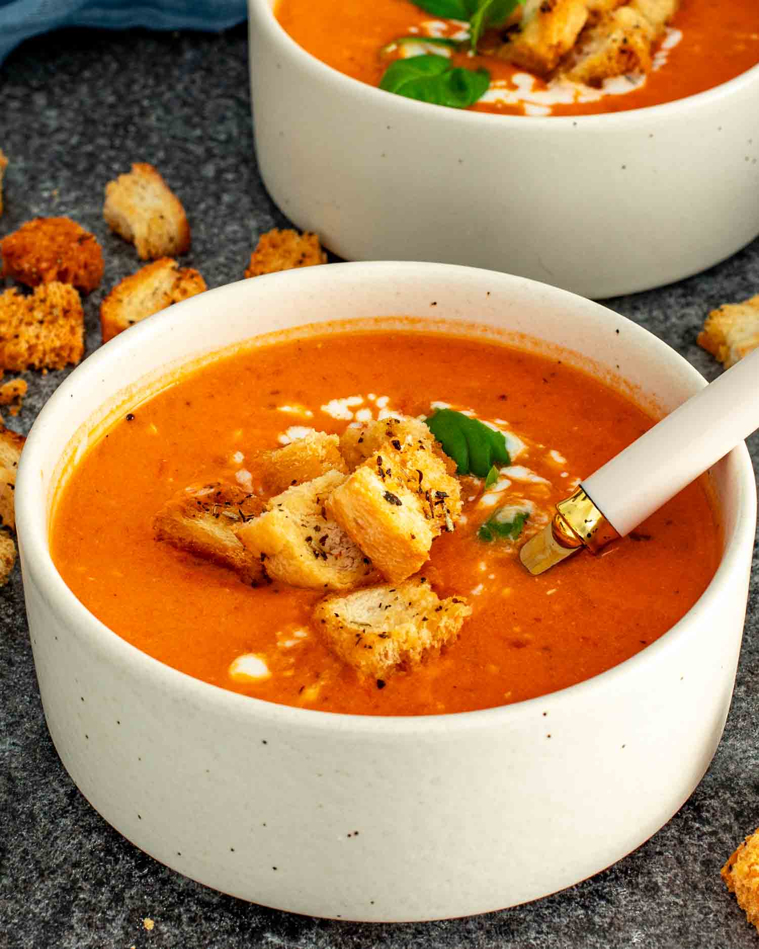 2 white speckled bowls of creamy tomato soup topped with golden croutons and fresh basil with a gold and white spoon resting in the front bowl surrounded by scattered croutons.