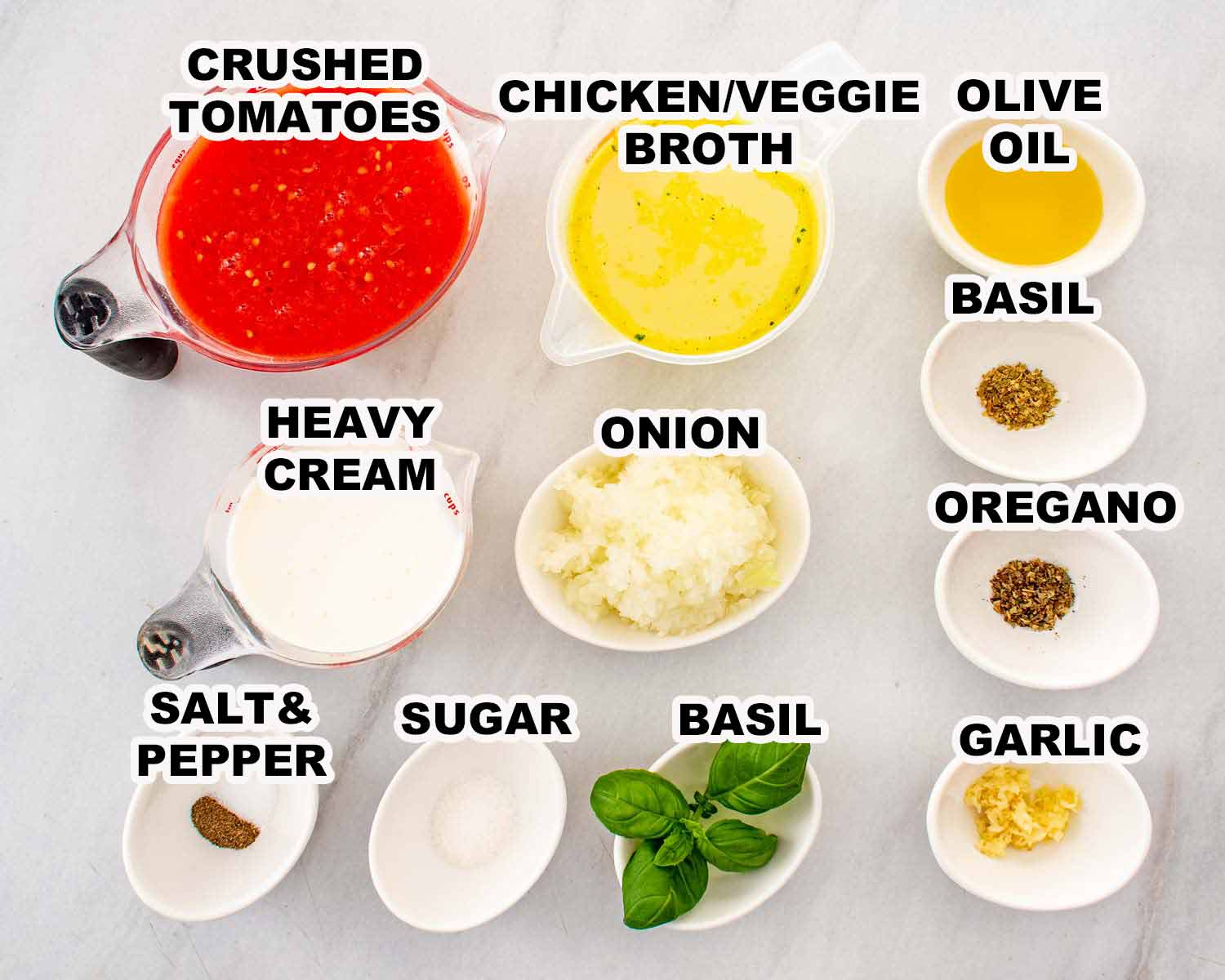 All ingredients for creamy tomato soup laid out on a white marble surface including crushed tomatoes, chicken broth, heavy cream, onion, garlic, olive oil, dried herbs, basil, salt and pepper.