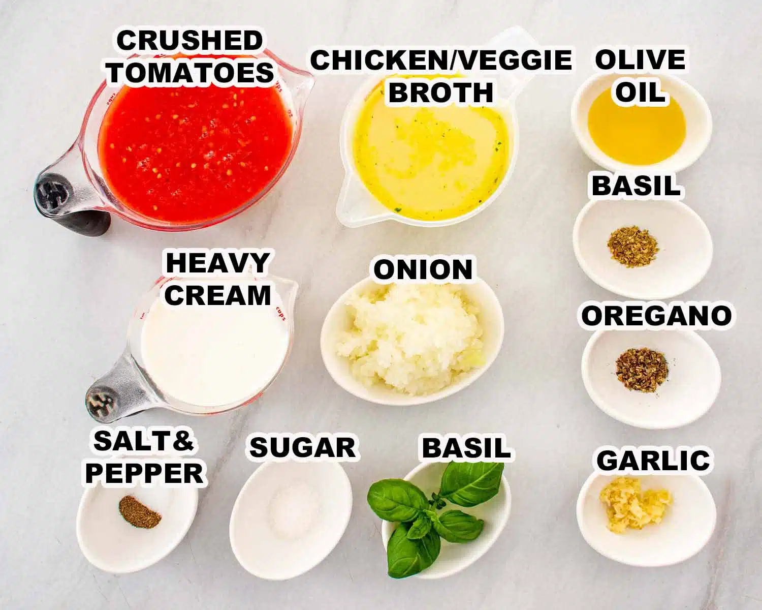 All ingredients for creamy tomato soup laid out on a white marble surface including crushed tomatoes, chicken broth, heavy cream, onion, garlic, olive oil, dried herbs, basil, salt and pepper.
