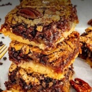 Elegant Pinterest pin for chocolate chip pecan pie bars showing three stacked bars on parchment paper with a gooey pecan chocolate filling and buttery shortbread base, with script text overlay reading Chocolate Chip Pecan Pie Bars.