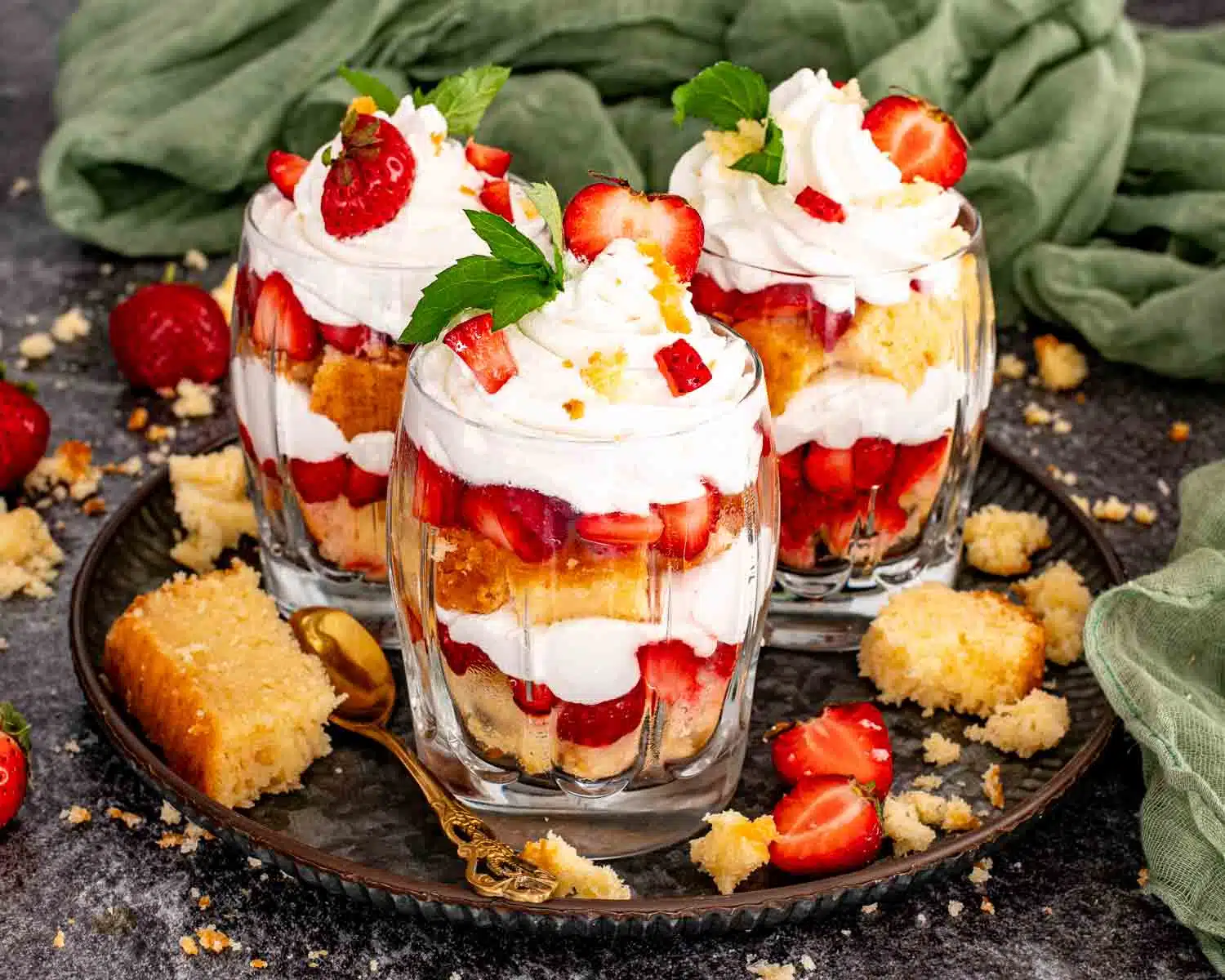 Three strawberry shortcake trifles on a dark plate showing generous layers of pound cake, macerated strawberries, and whipped cream topped with fresh strawberries, mint, and pound cake crumbles.
