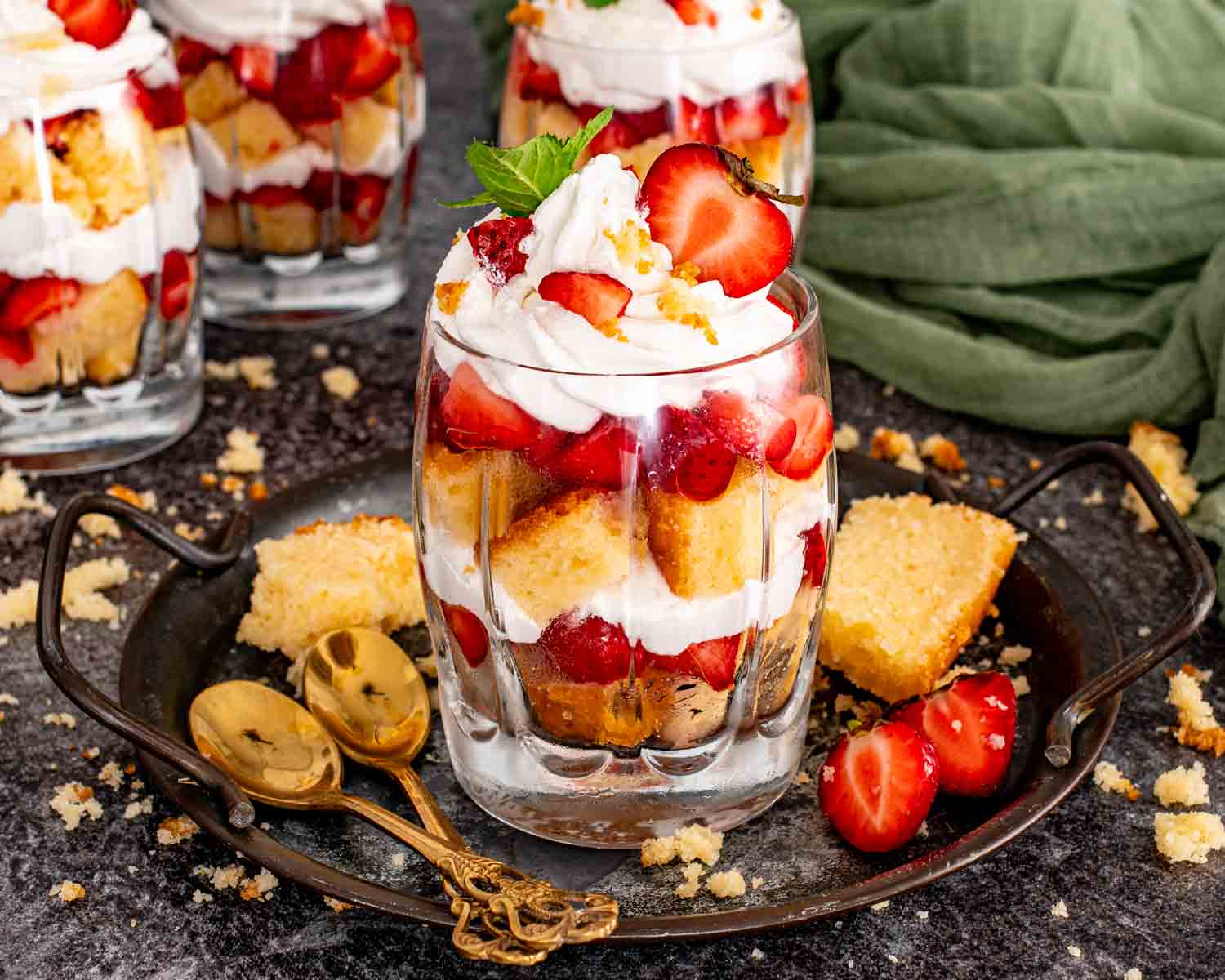 A single strawberry shortcake trifle on a dark metal tray showing layered pound cake, fresh strawberries, and whipped cream topped with a halved strawberry, mint, and two gold spoons alongside.