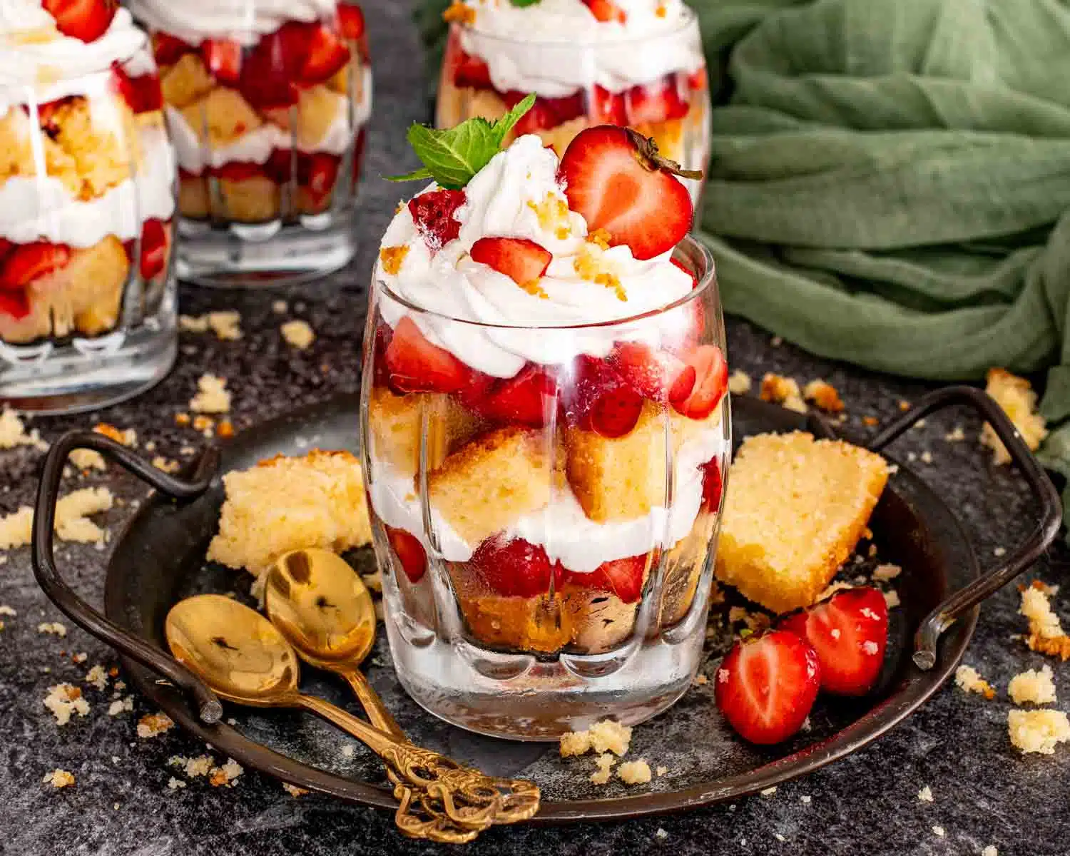 A single strawberry shortcake trifle on a dark metal tray showing layered pound cake, fresh strawberries, and whipped cream topped with a halved strawberry, mint, and two gold spoons alongside.