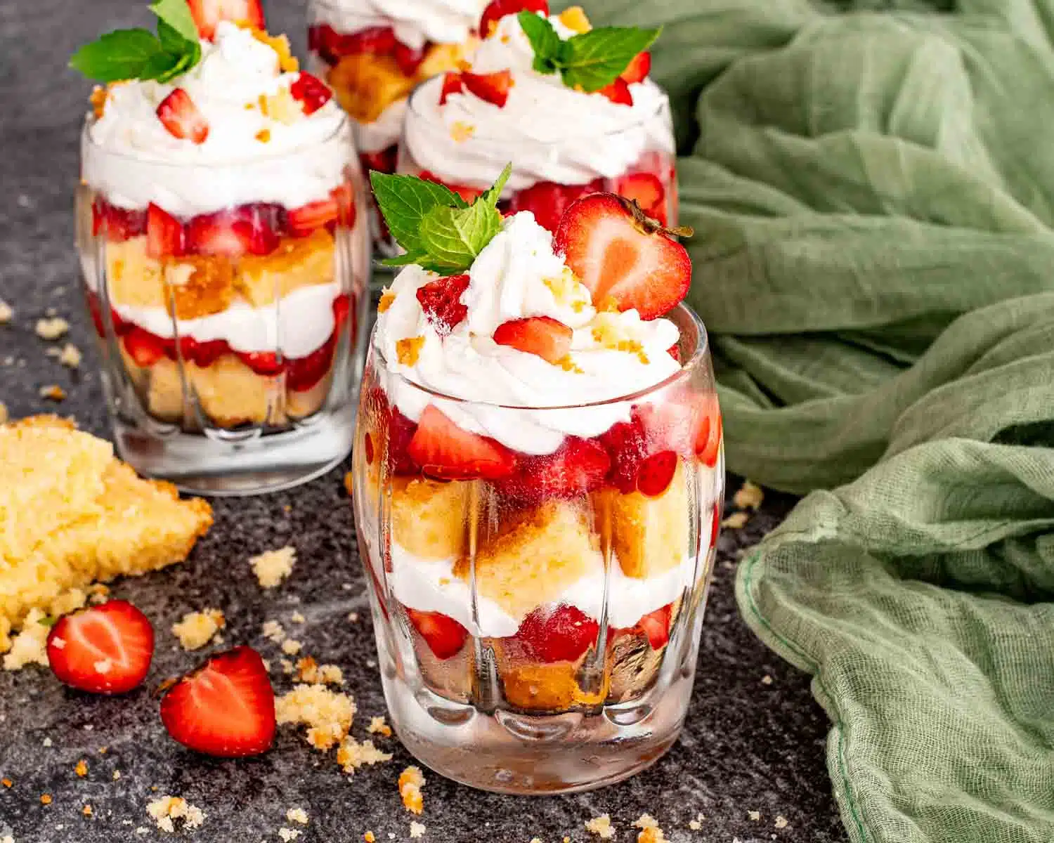 Three strawberry shortcake trifles in clear glasses showing layers of pound cake, macerated strawberries, and billowy whipped cream topped with fresh strawberries, mint, and pound cake crumbles.