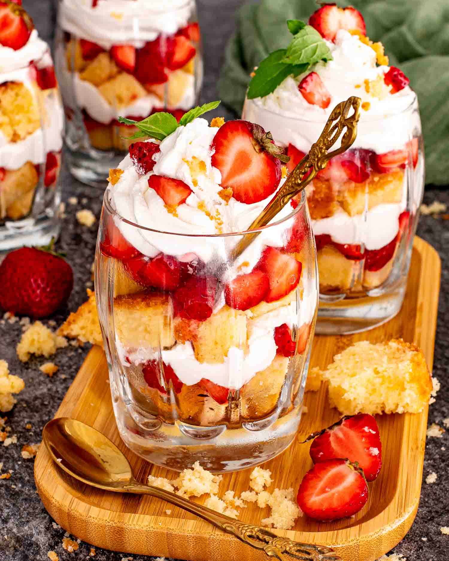 Three strawberry shortcake trifles in clear glasses on a wood board showing layered pound cake, macerated strawberries, and whipped cream topped with fresh strawberries, mint, and a gold spoon.