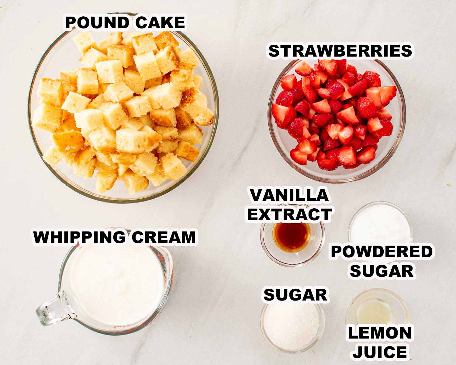 Overhead flat lay of strawberry shortcake trifle ingredients including pound cake cubes, fresh strawberries, whipping cream, vanilla extract, powdered sugar, granulated sugar, and lemon juice.