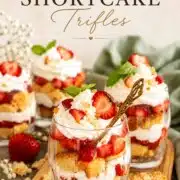 Pinterest pin of strawberry shortcake trifles in clear glasses on a wood board with whipped cream, macerated strawberries, and pound cake layers with elegant script text overlay reading Strawberry Shortcake Trifles.
