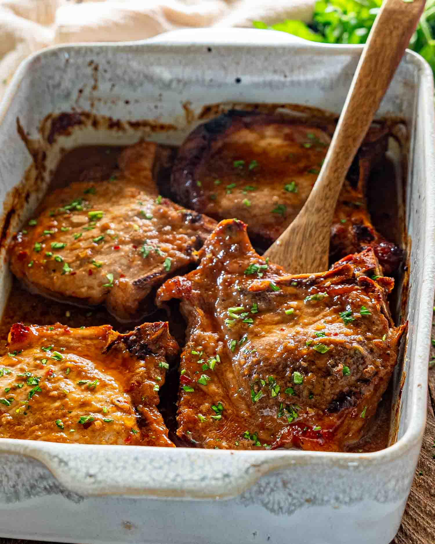 Wooden spoon lifting a tender glazed pork chop from baking dish filled with flavorful brown sugar sauce.