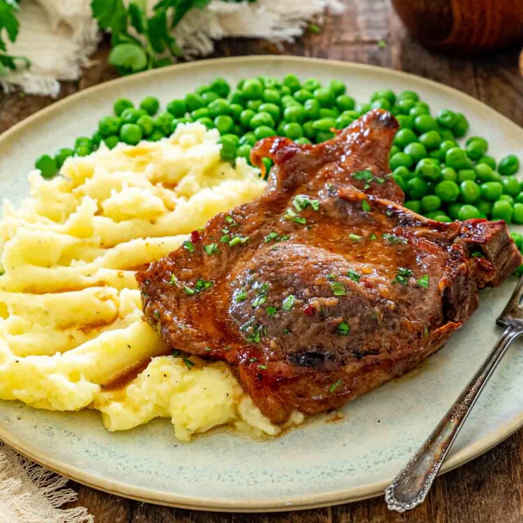 Glazed bone-in pork chop served whole with creamy mashed potatoes and green peas on a rustic plate.