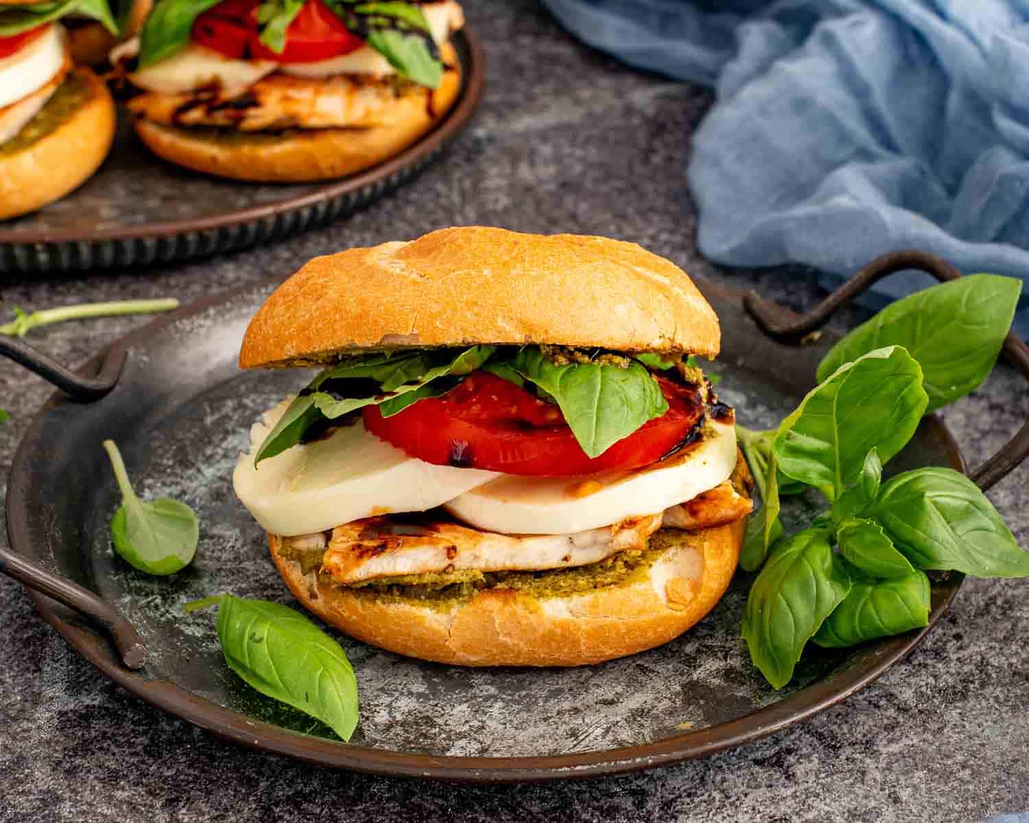 Grilled chicken Caprese sandwich on a rustic serving plate with creamy mozzarella, ripe tomatoes, basil, pesto, and balsamic glaze.