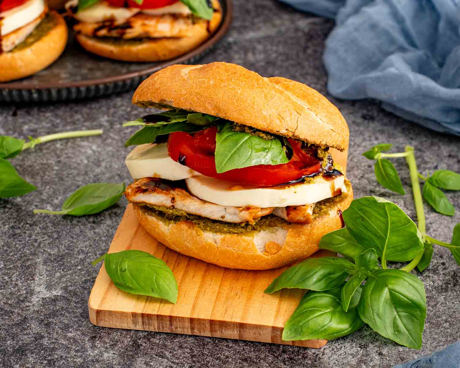 Grilled chicken Caprese sandwich served on a wooden board with fresh basil, mozzarella, tomatoes, pesto, and balsamic glaze.