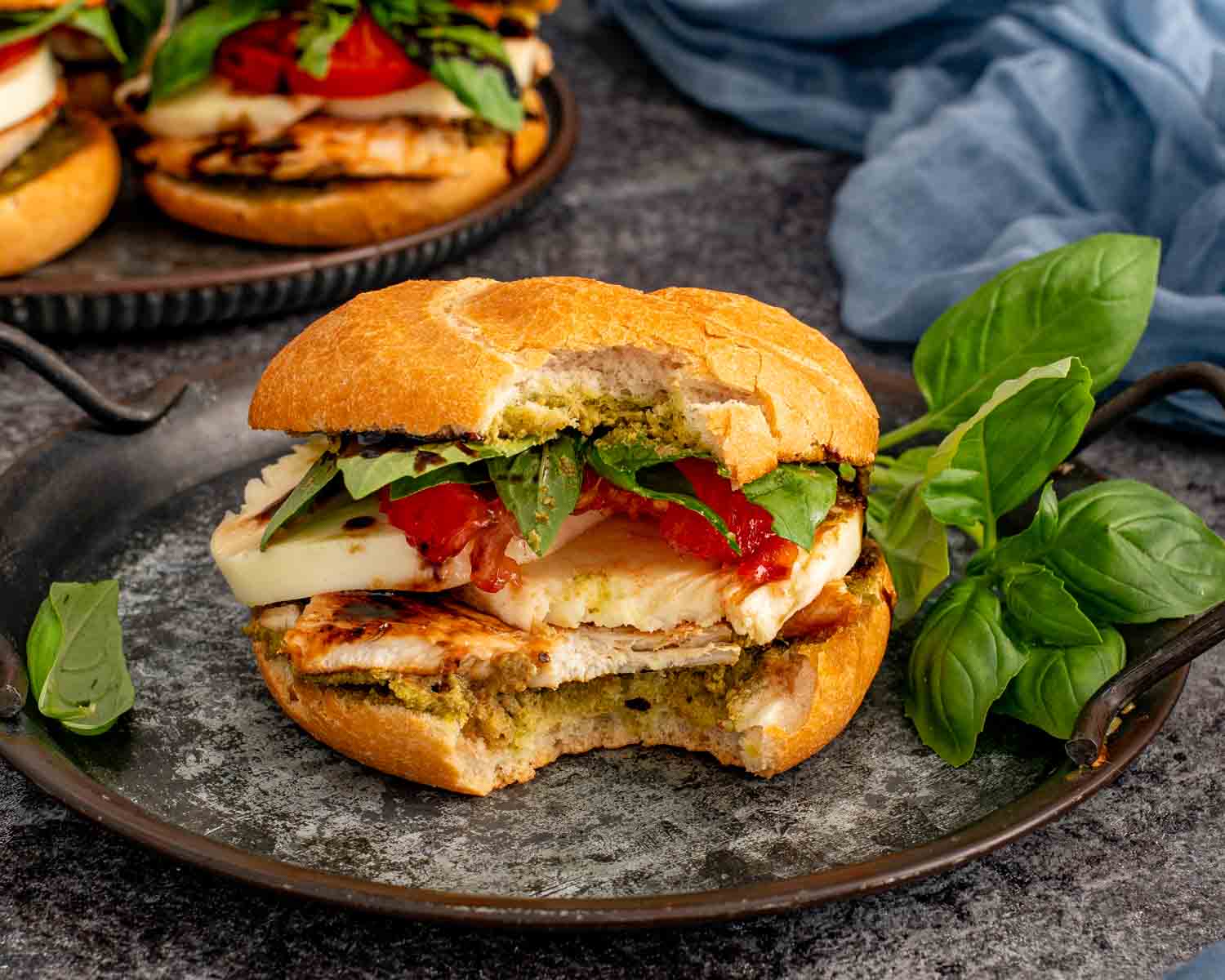 Grilled chicken Caprese sandwich with a bite taken out, revealing juicy chicken, melted mozzarella, pesto, tomatoes, and fresh basil.