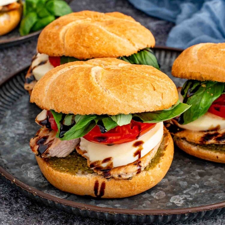 Three grilled chicken Caprese sandwiches on a serving platter layered with pesto, fresh mozzarella, tomatoes, basil, and balsamic glaze.
