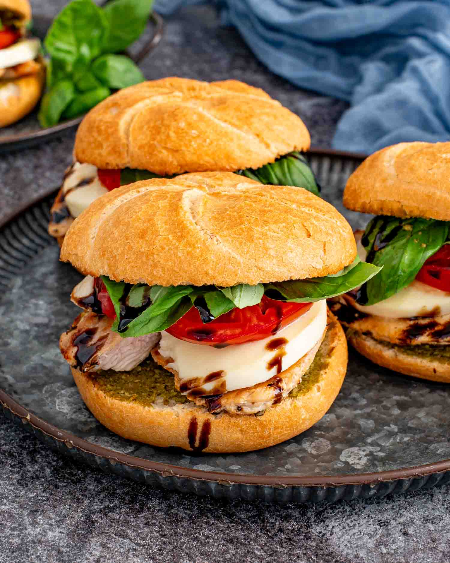 Three grilled chicken Caprese sandwiches on a serving platter layered with pesto, fresh mozzarella, tomatoes, basil, and balsamic glaze.