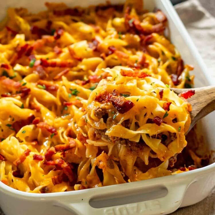 Spoonful of cheesy beef casserole lifted from baking dish showing creamy layers, noodles, bacon, and melted cheese.