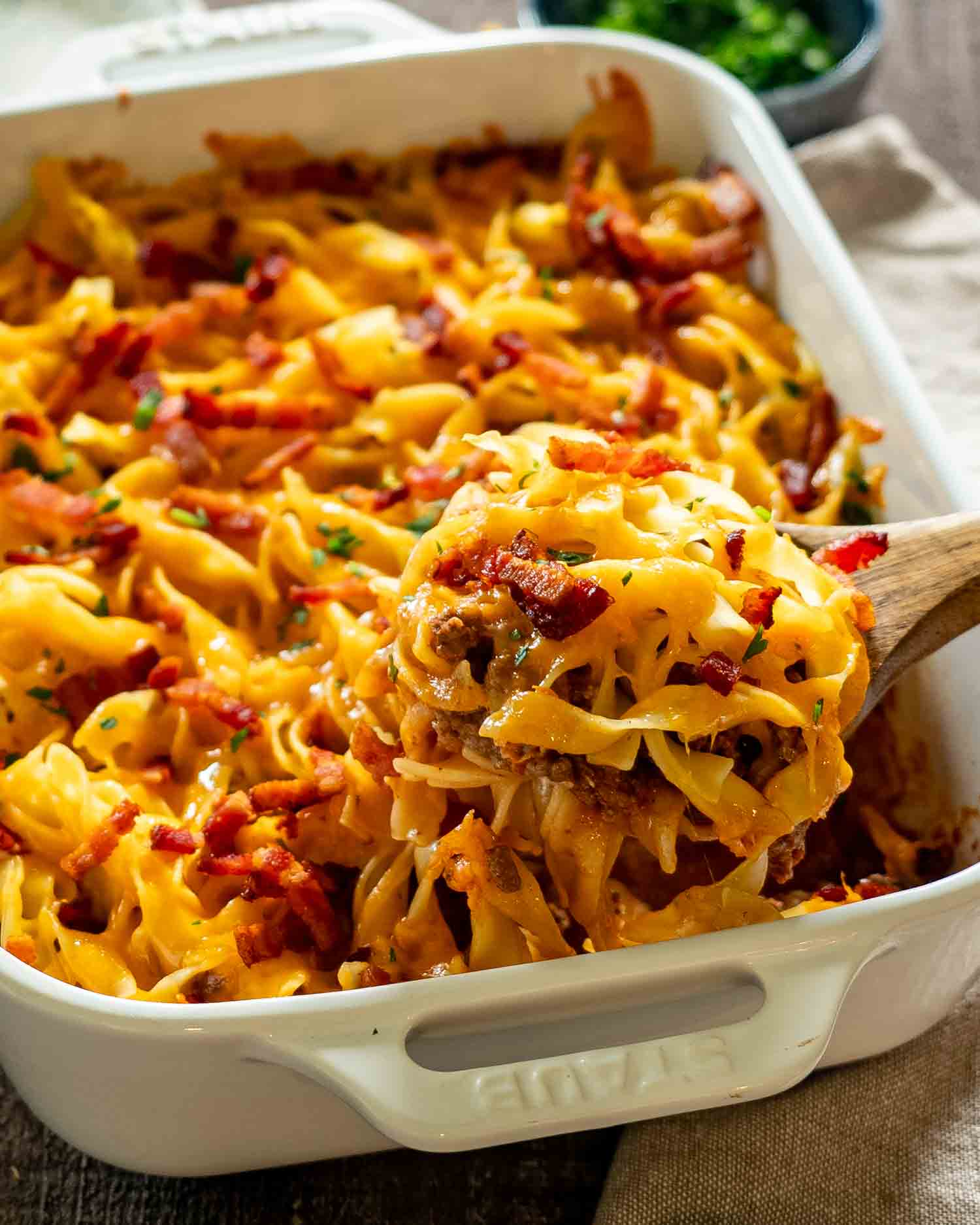 Spoonful of cheesy beef casserole lifted from baking dish showing creamy layers, noodles, bacon, and melted cheese.