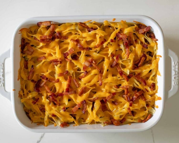 Baked million dollar beef casserole topped with melted cheddar cheese and crispy bacon in white ceramic baking dish.
