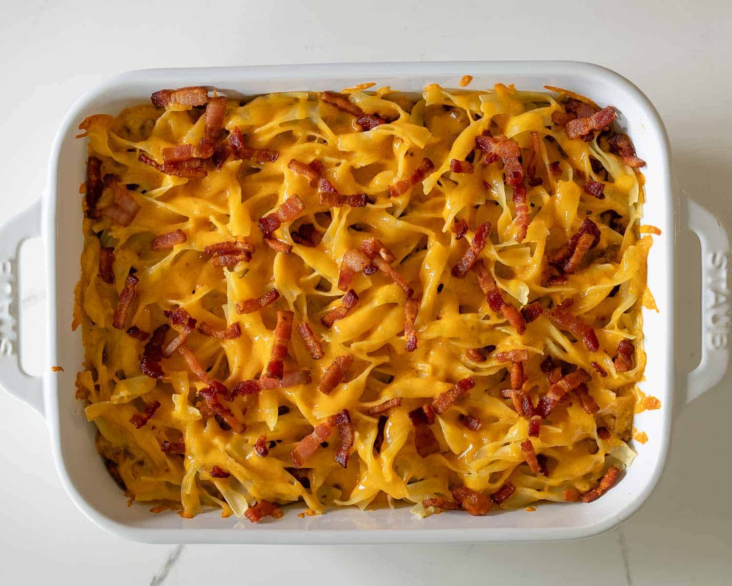 Baked million dollar beef casserole topped with melted cheddar cheese and crispy bacon in white ceramic baking dish.