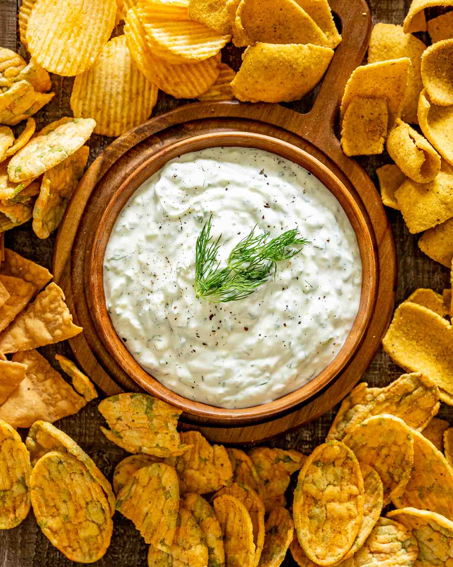 Overhead view of creamy tzatziki garnished with fresh dill in a wooden bowl, surrounded by assorted golden vegetable chips.