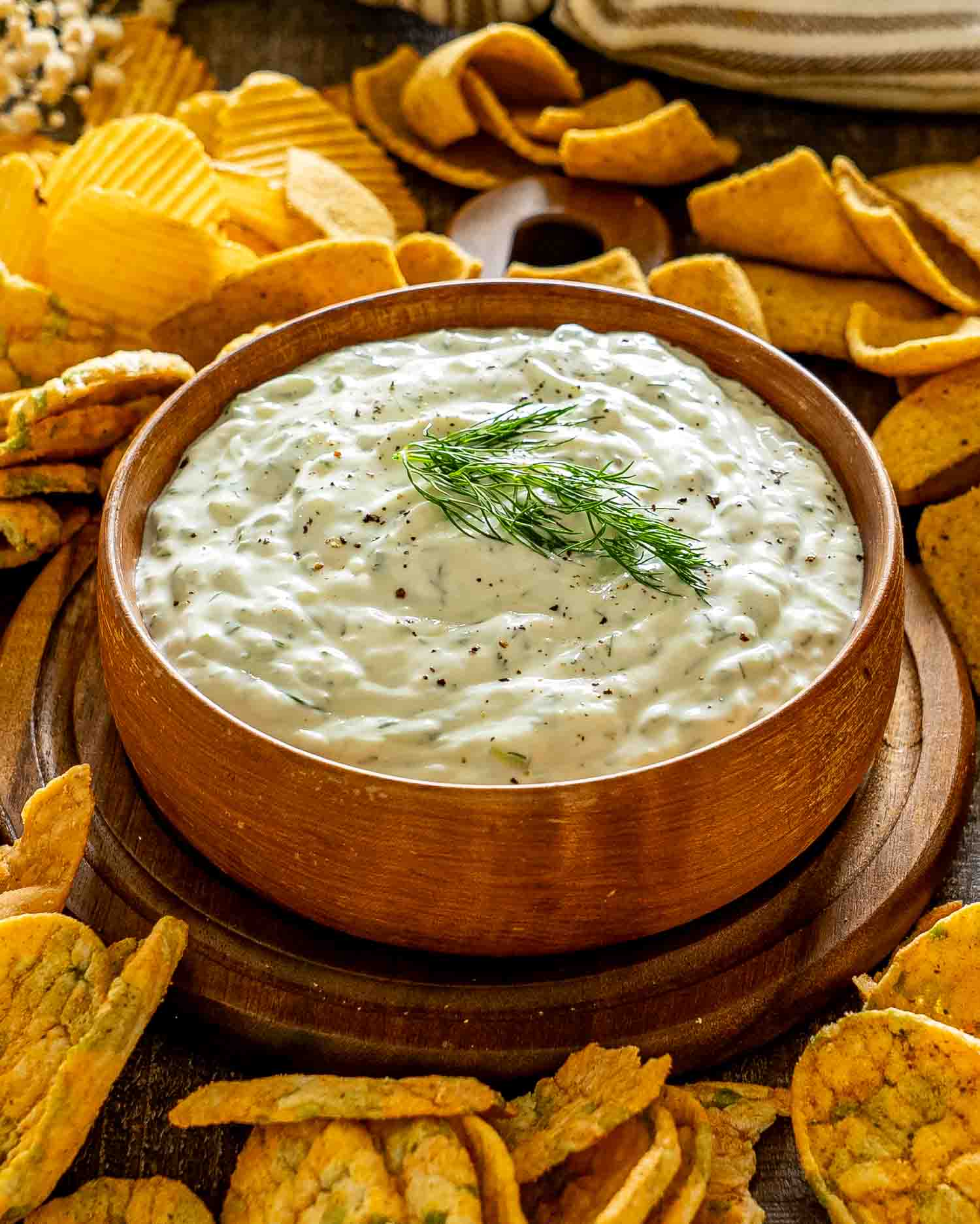 Rustic wooden bowl filled with creamy dill tzatziki sauce, surrounded by an assortment of golden, crunchy vegetable chips.