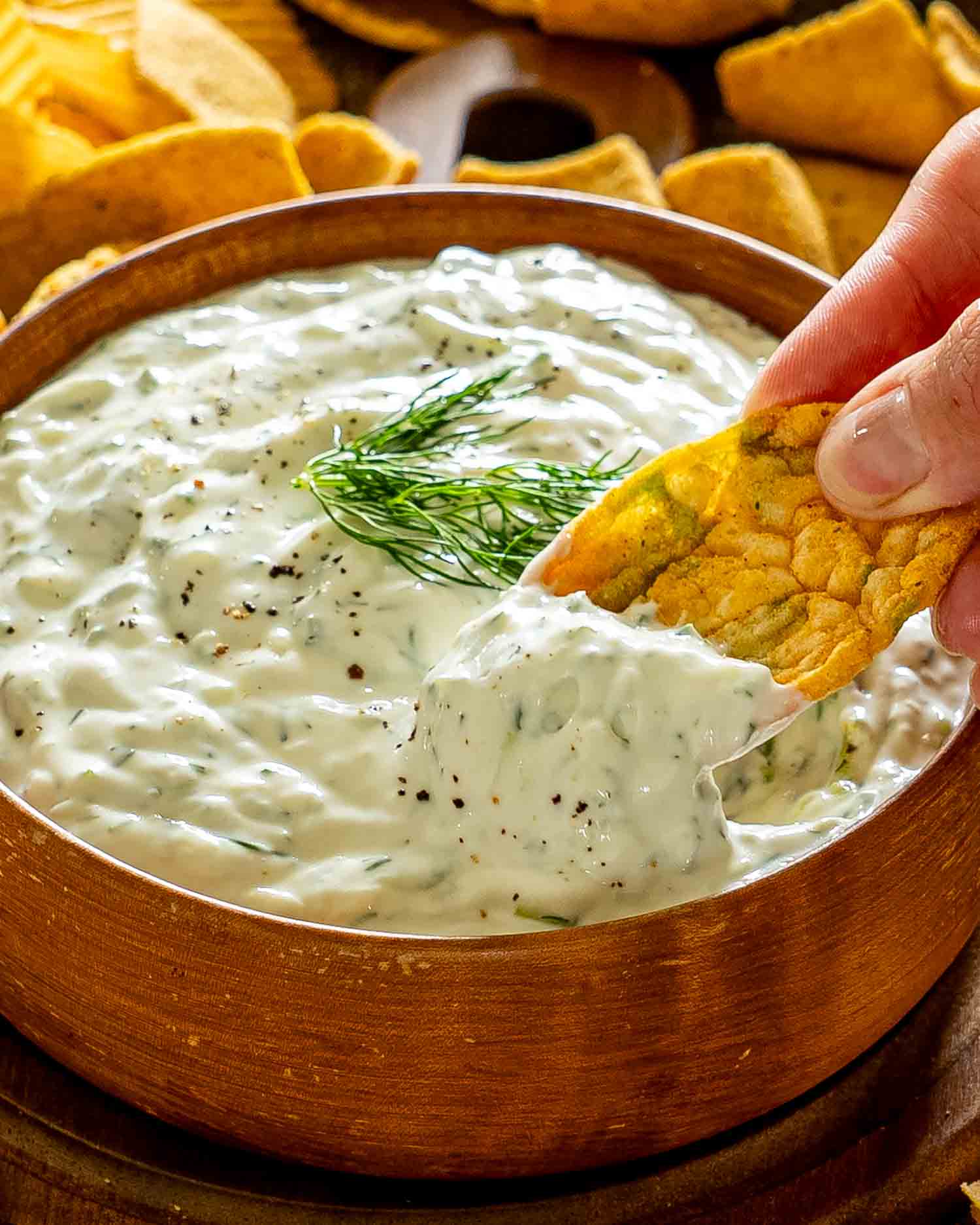 A crisp chip scoops thick, creamy tzatziki garnished with fresh dill from a rustic wooden serving bowl.