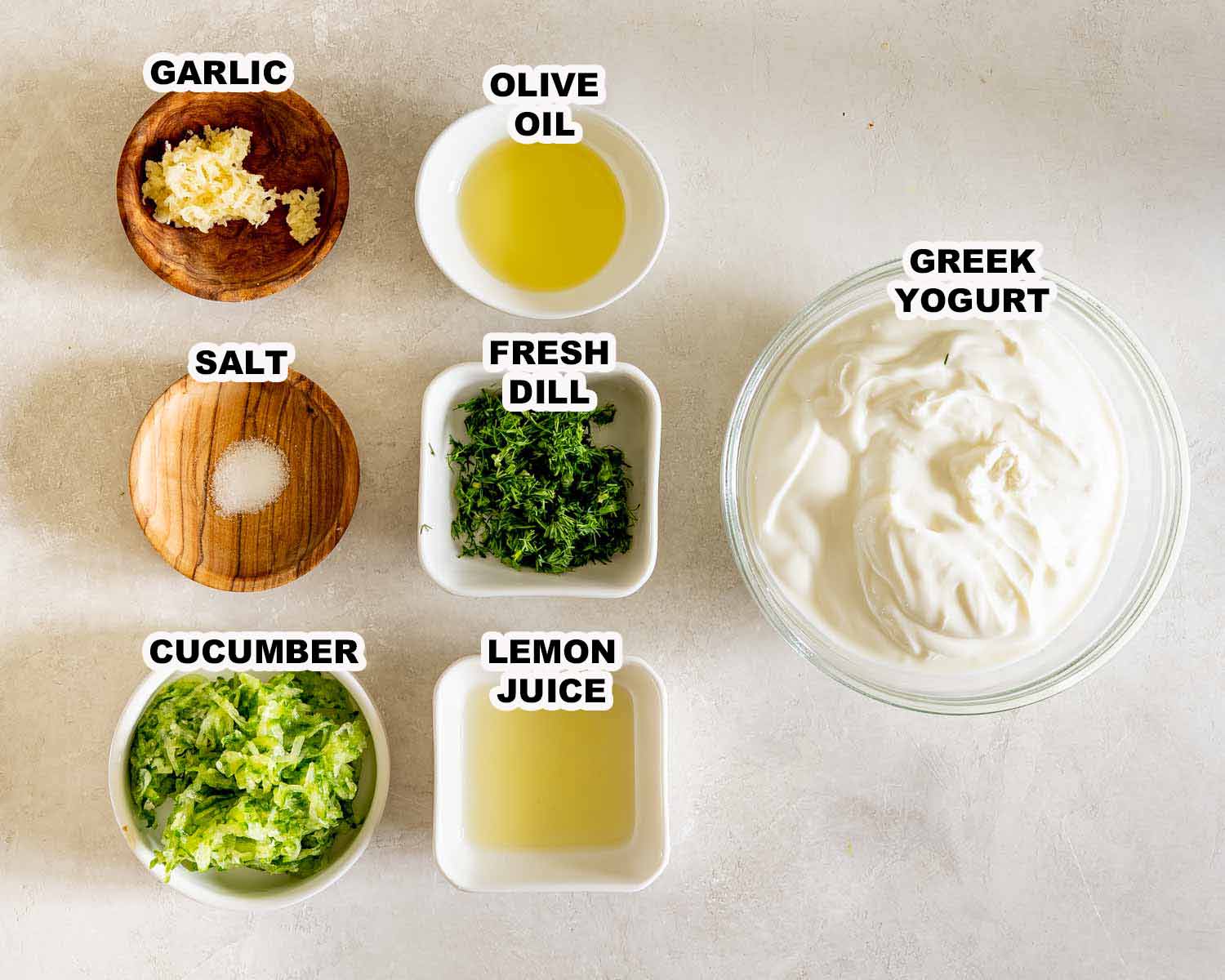 Overhead arrangement of labeled tzatziki ingredients including Greek yogurt, cucumber, dill, garlic, olive oil, lemon juice, and salt.