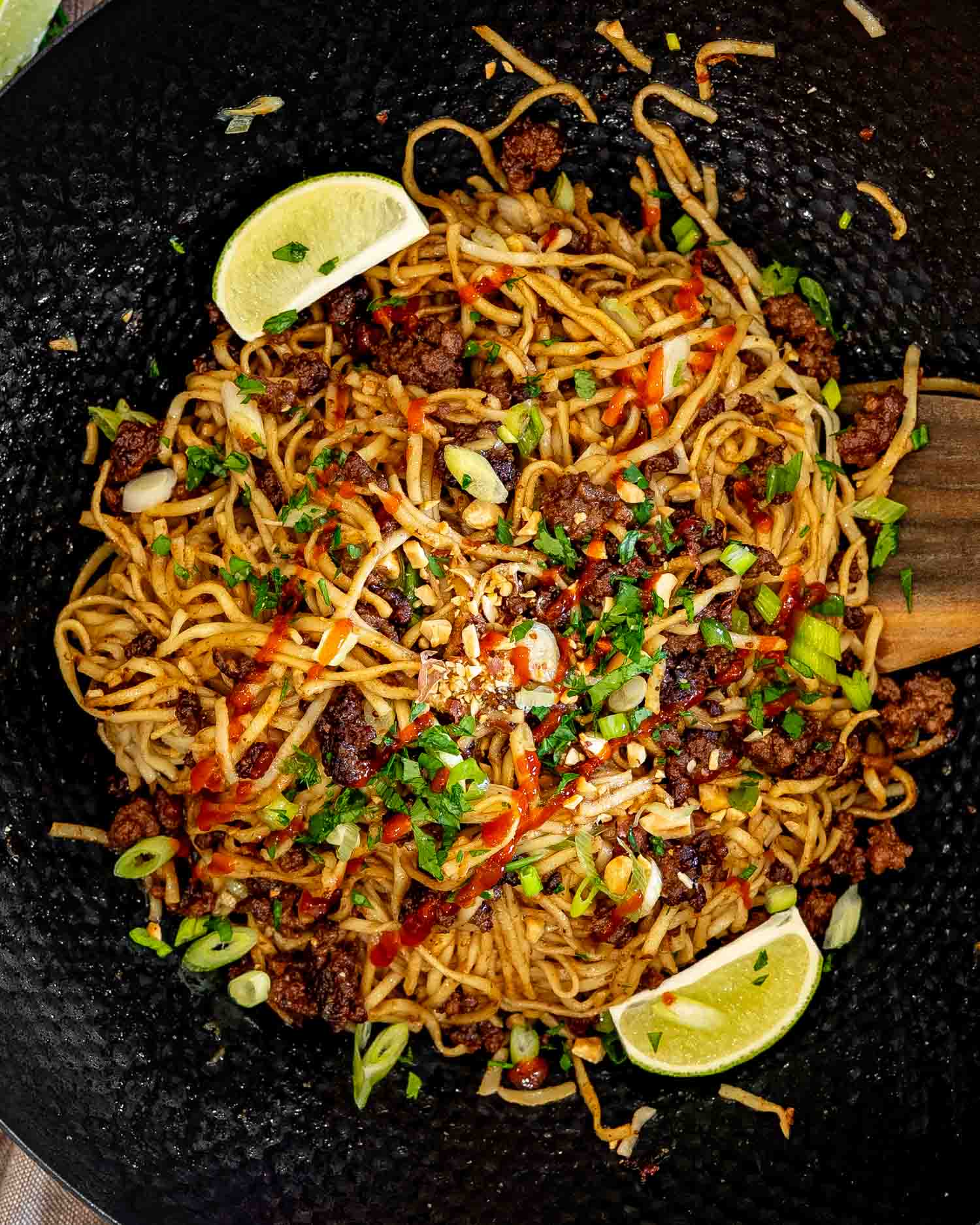 Overhead view of caramelized beef and peanut noodles in a wok, garnished with cilantro, peanuts, green onions, sriracha, and lime.