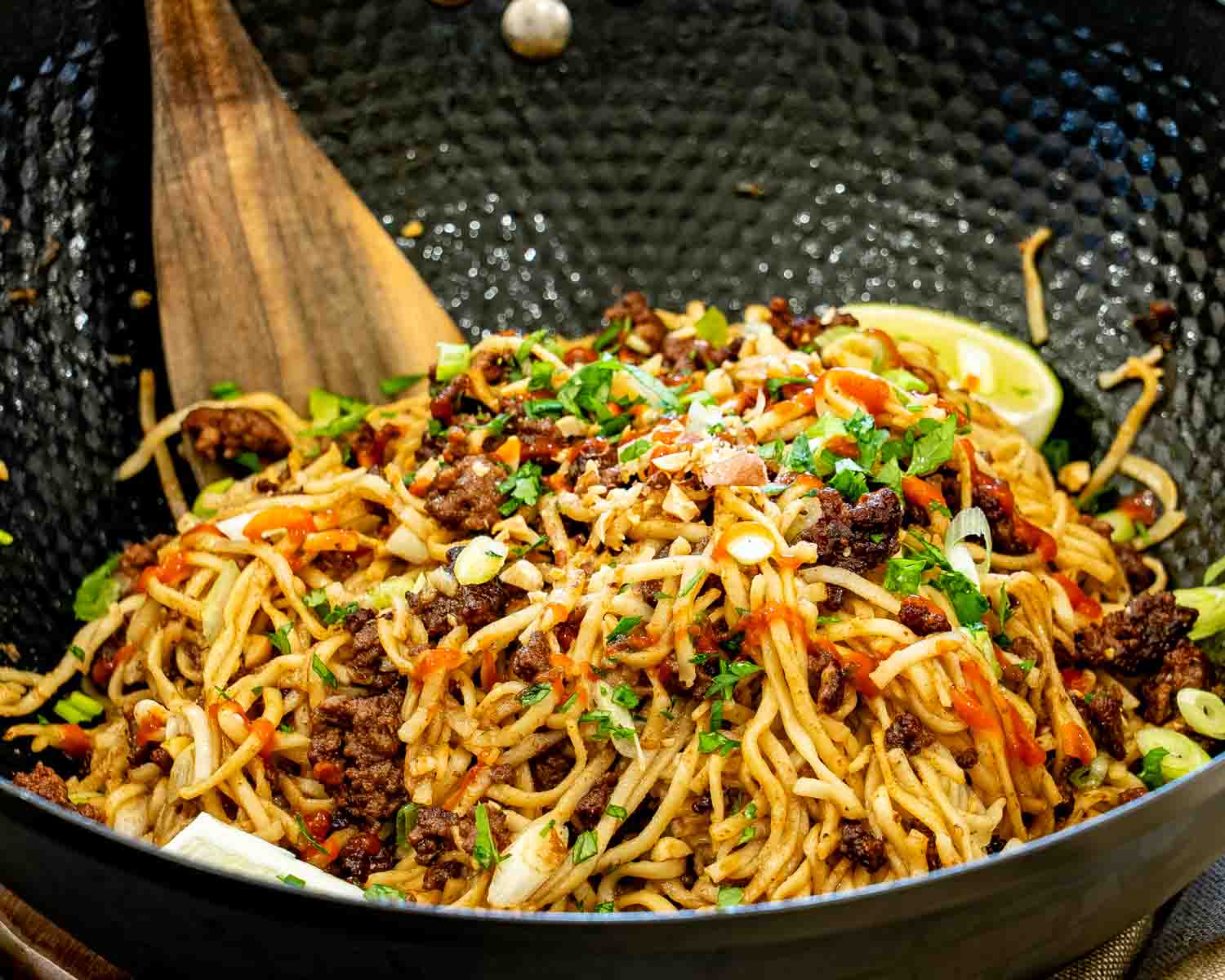 Close-up of caramelized beef and peanut noodles in a black wok, topped with chopped peanuts, cilantro, green onions, and sriracha.