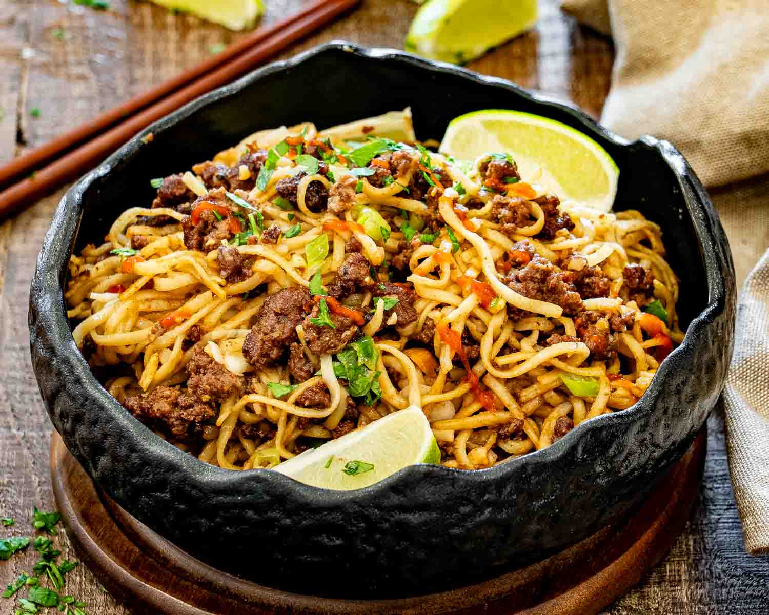 Black bowl filled with caramelized beef and peanut noodles, garnished with cilantro, green onions, sriracha, peanuts, and lime wedges.