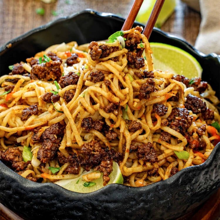 Close-up of glossy caramelized beef and peanut noodles lifted with chopsticks, served in a black bowl with lime wedges.