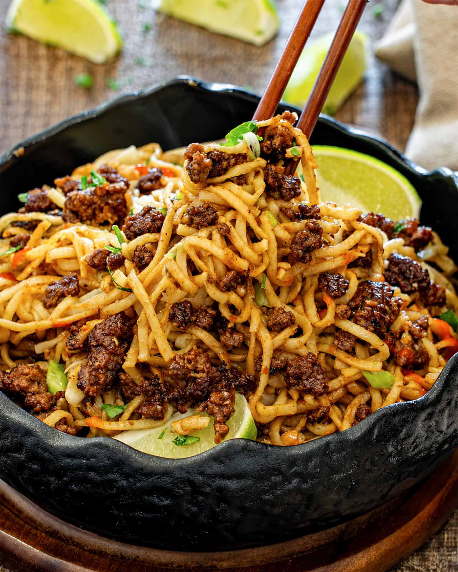 Close-up of glossy caramelized beef and peanut noodles lifted with chopsticks, served in a black bowl with lime wedges.