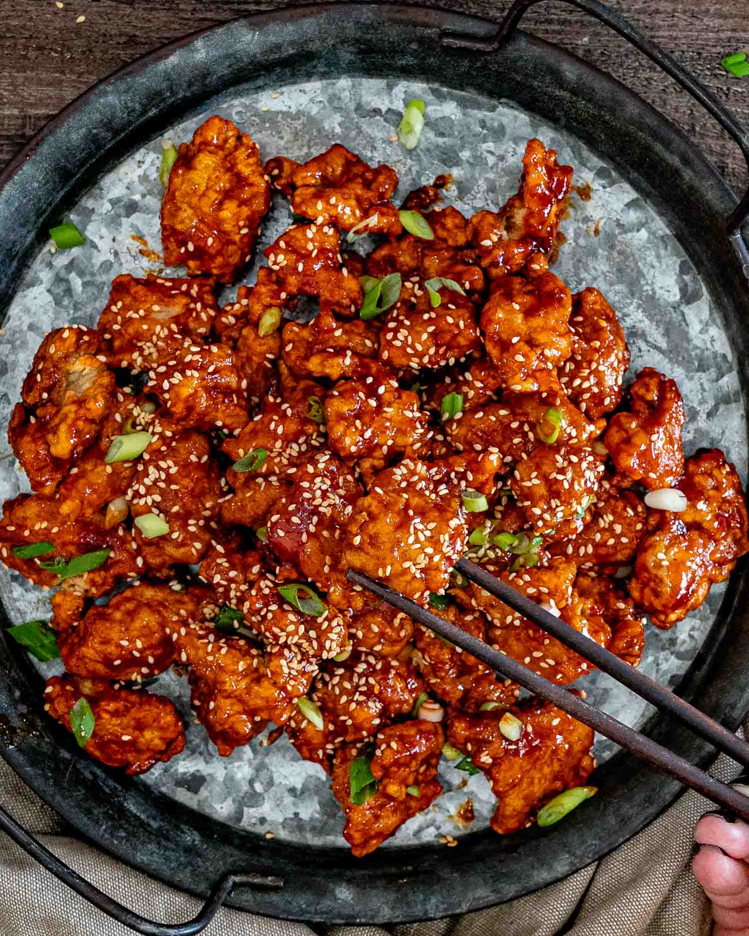Overhead view of glossy Korean popcorn chicken on a dark platter, garnished with sesame seeds, green onions, and chopsticks.