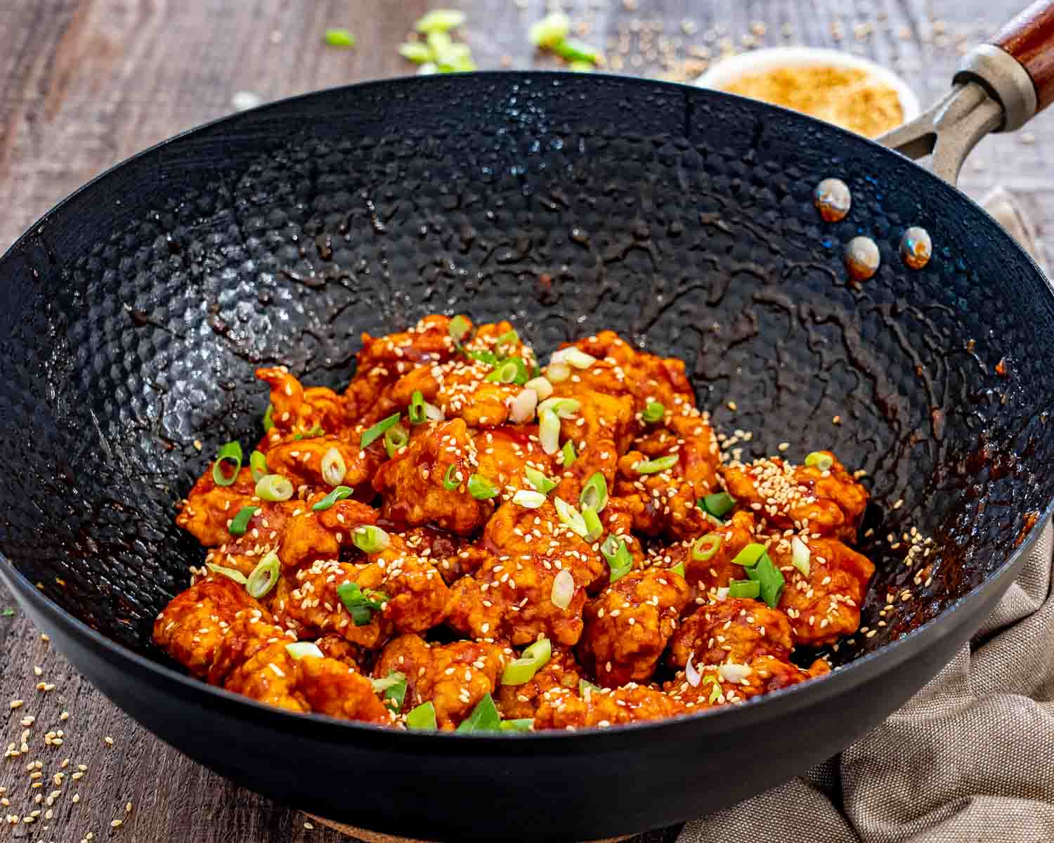 Saucy Korean popcorn chicken rests in a black wok, finished with toasted sesame seeds and plenty of sliced green onions.