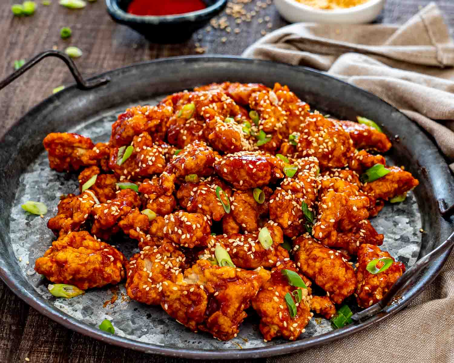 Finished Korean popcorn chicken fills a dark platter, glistening with spicy sauce and scattered sesame seeds and sliced green onions.