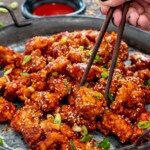 Chopsticks lift one glossy piece of Korean popcorn chicken from a platter garnished with sesame seeds and green onions.