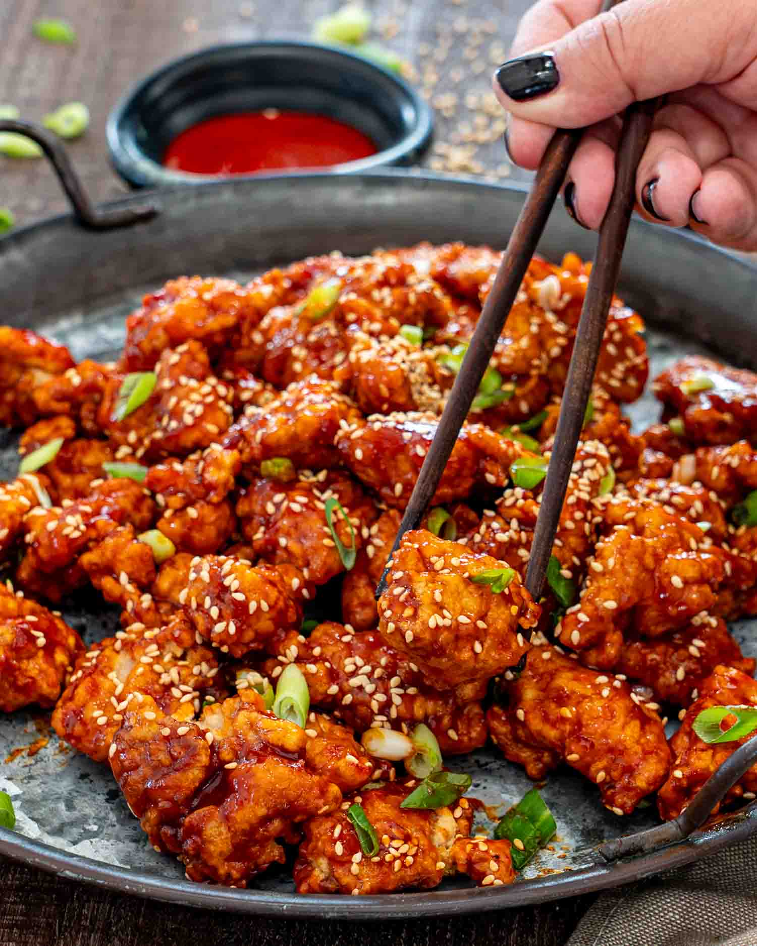 Chopsticks lift one glossy piece of Korean popcorn chicken from a platter garnished with sesame seeds and green onions.