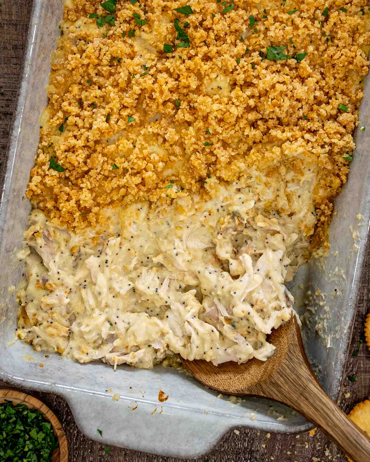 Wooden spoon serves creamy poppy seed chicken casserole from a baking dish topped with golden crumbs and parsley.