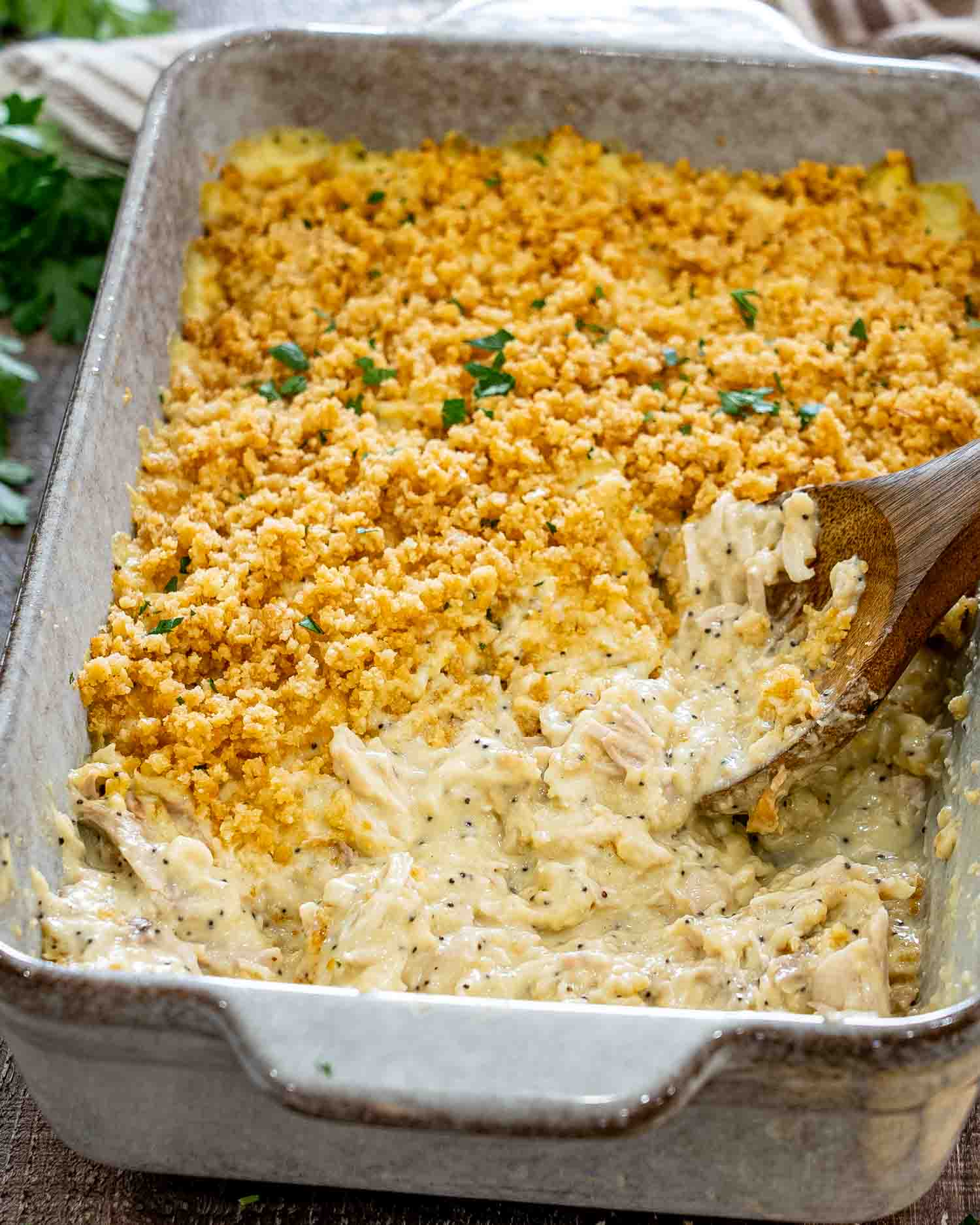 Close-up of creamy poppy seed chicken casserole with a golden cracker topping, fresh parsley, and wooden serving spoon.