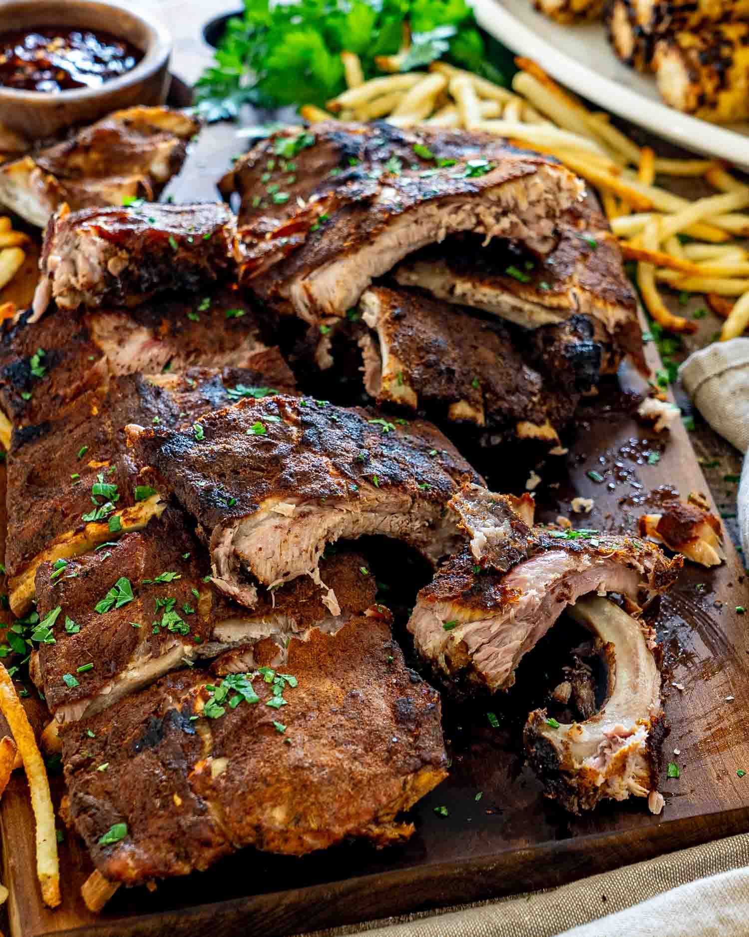 Sliced Memphis-style pork ribs piled on a wooden serving board with fries, barbecue sauce, and parsley.