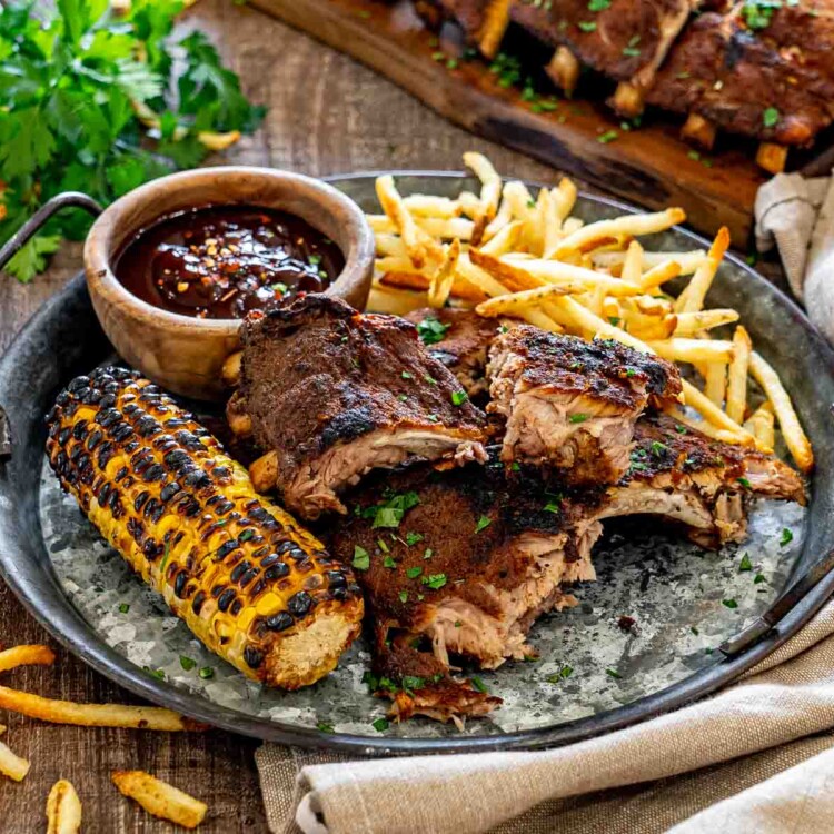 Memphis-style pork ribs served with grilled corn, crispy fries, barbecue sauce, and fresh parsley on a rustic platter.