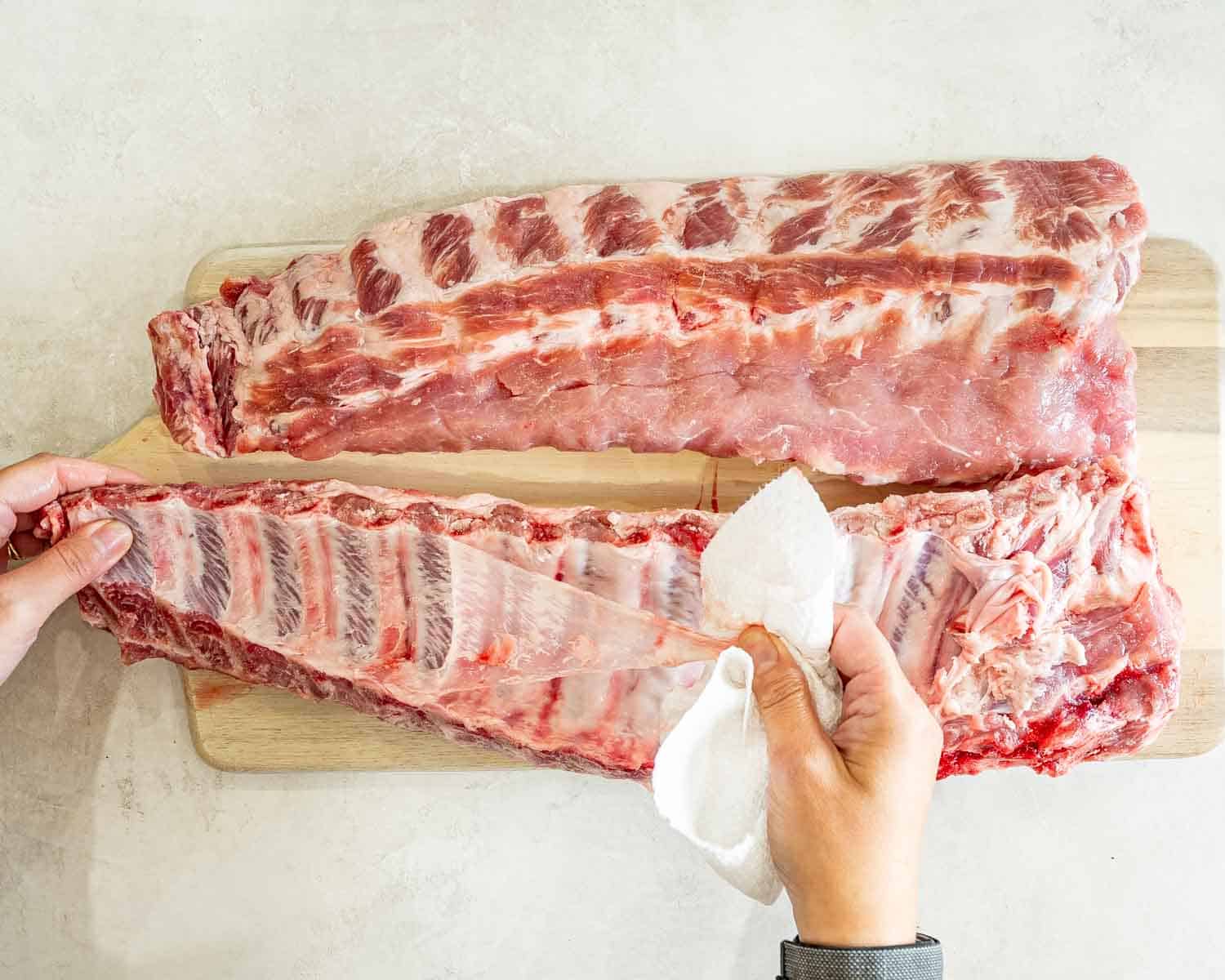 Hands remove the thin membrane from the back of raw pork ribs using a paper towel.