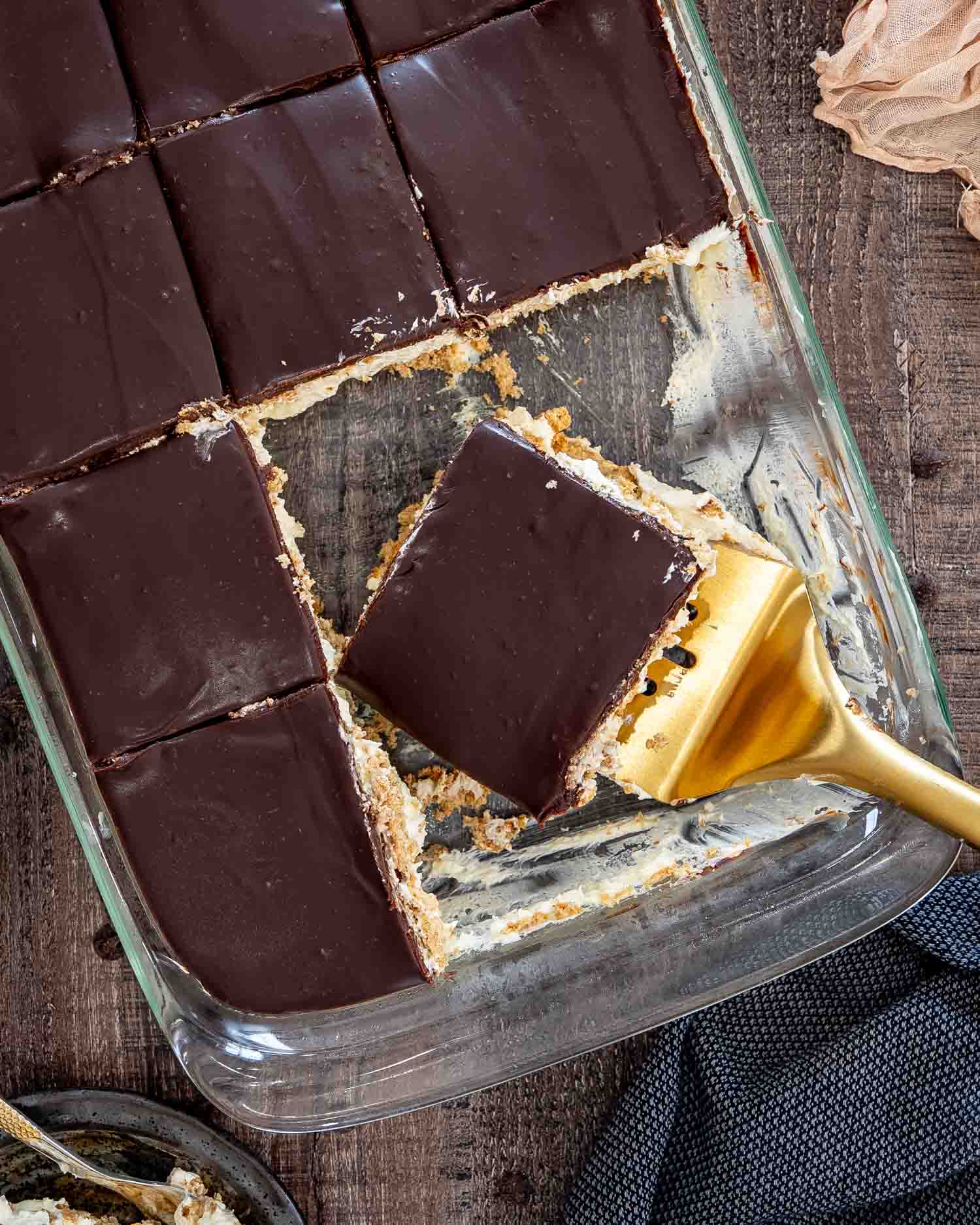 An overhead view shows chocolate eclair cake squares in a glass dish with one serving being lifted out.