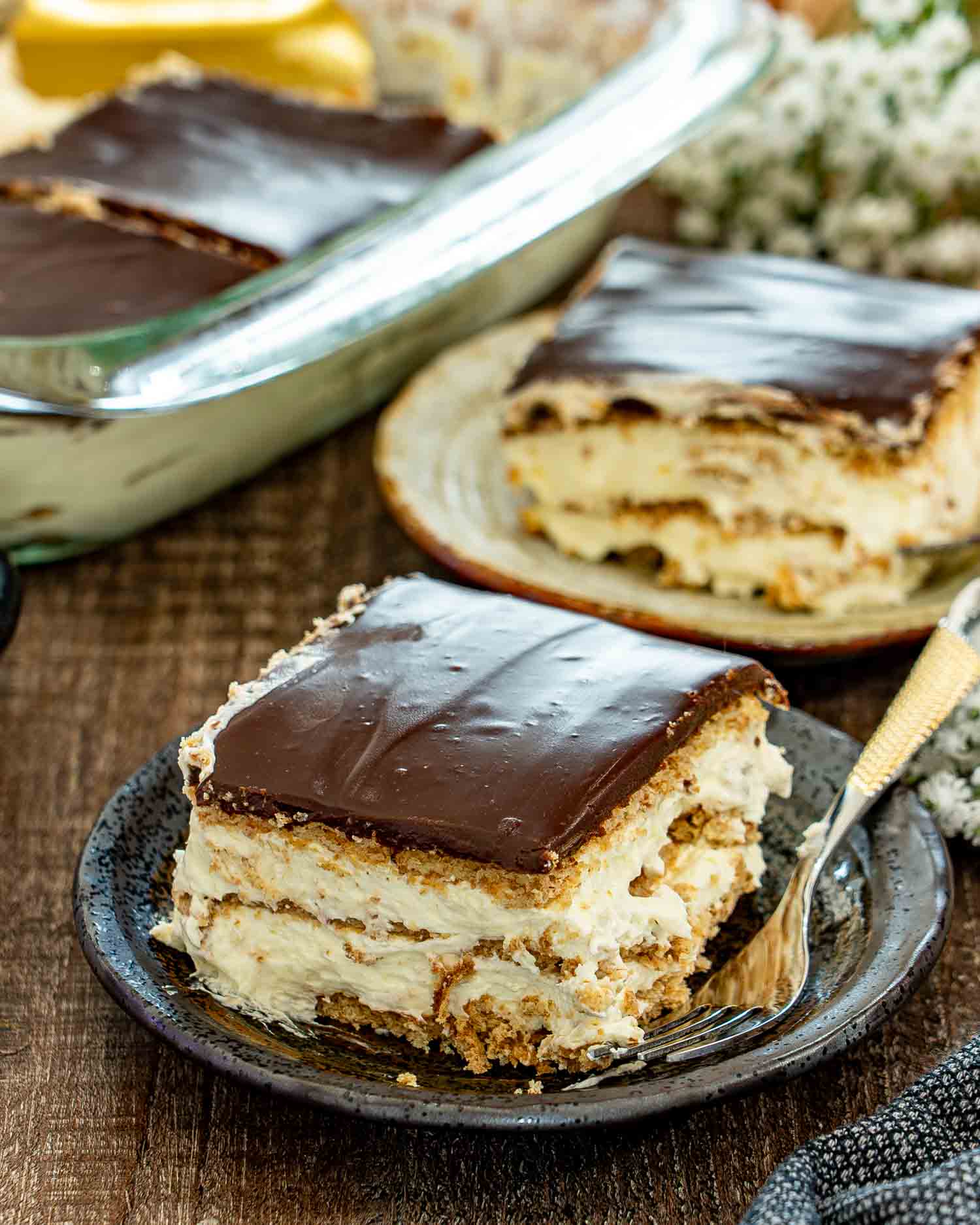 A thick slice of no bake chocolate eclair cake with creamy layers and ganache sits on a dark plate.