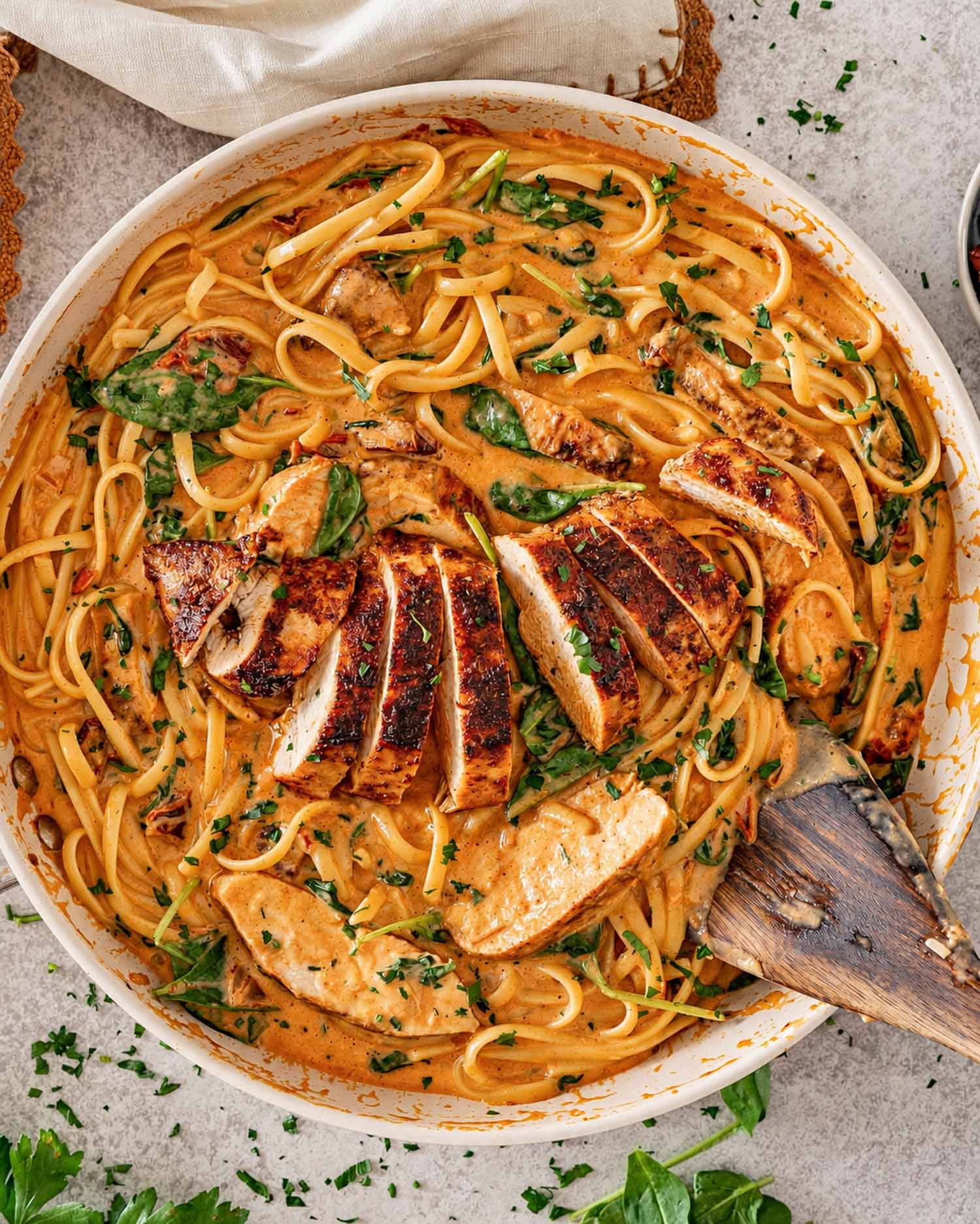Overhead view of sliced chicken resting on creamy sun-dried tomato linguine with spinach in a white skillet.