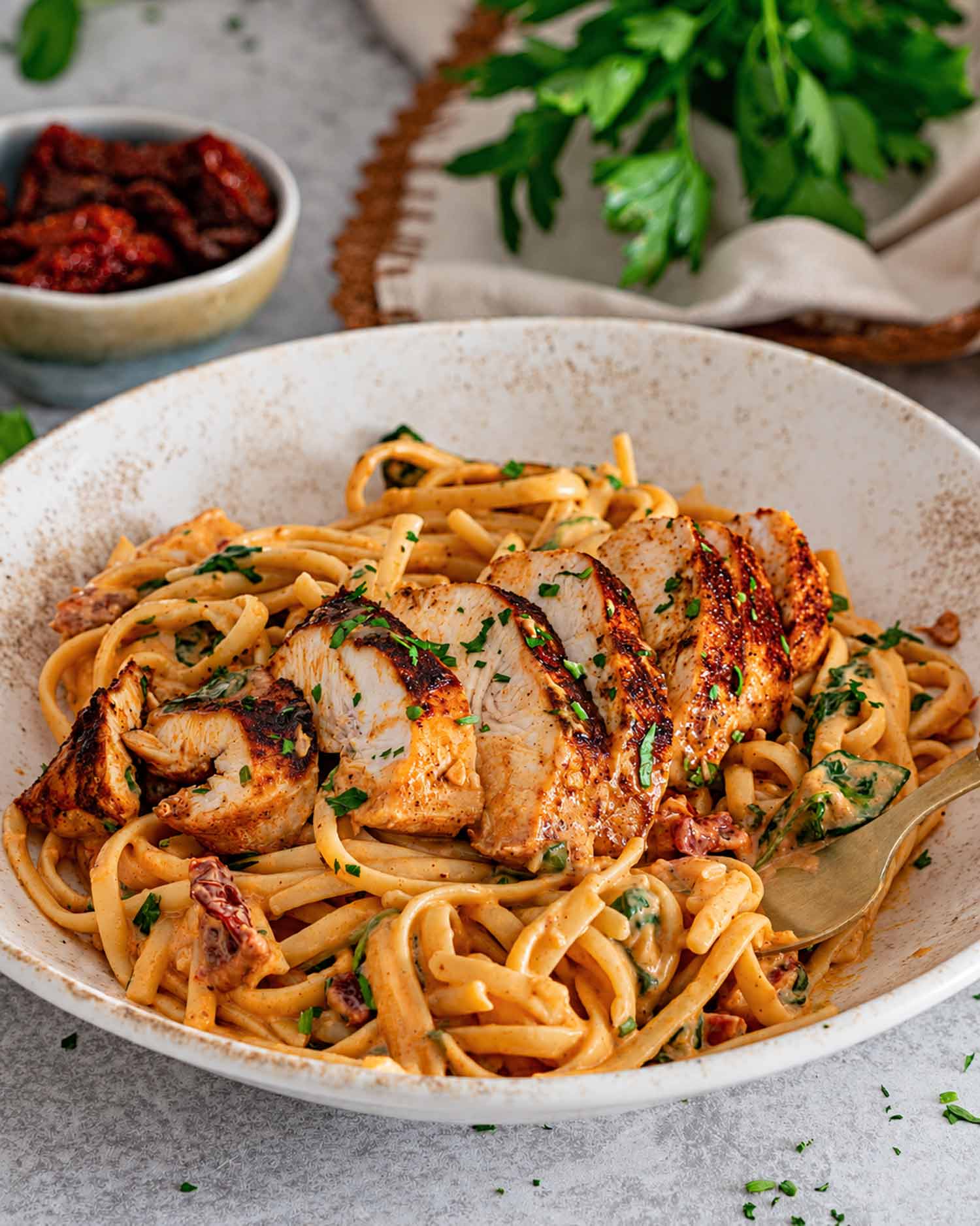 Close view of browned chicken slices on creamy sun-dried tomato linguine, with a fork tucked into the bowl.