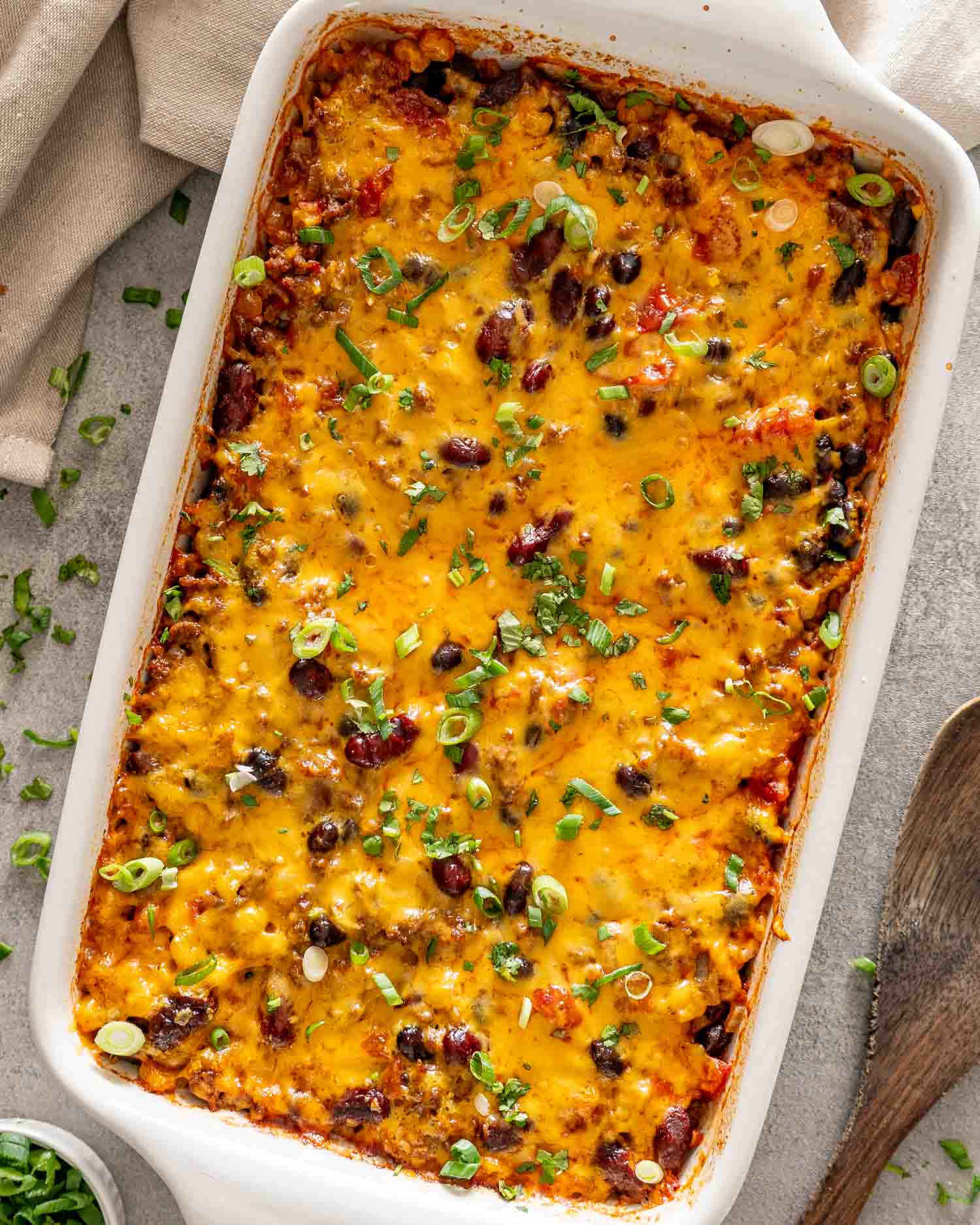 Overhead view of chili rice bake in a white casserole dish, topped with melted cheddar, sliced green onions, and cilantro.
